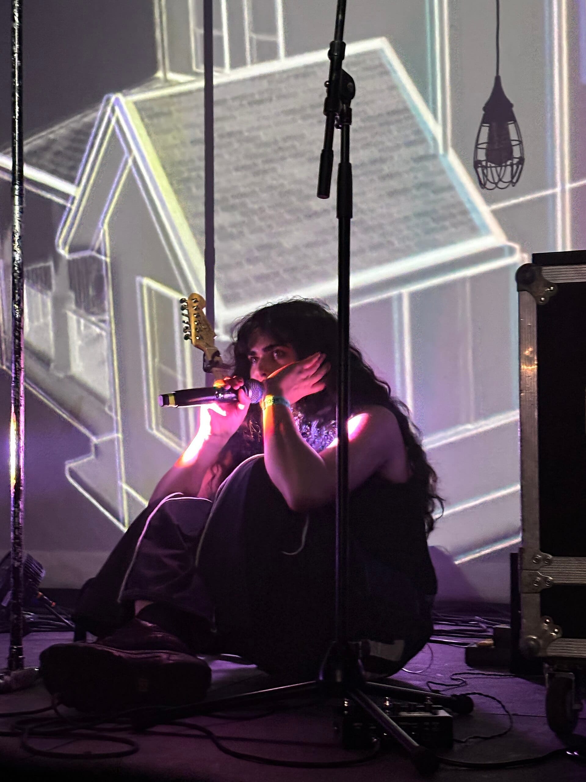 Underscores sits on the stage floor holding a microphone to her mouth, long dark hair loose, lit pink and purple with an outlined house projected on the wall behind.