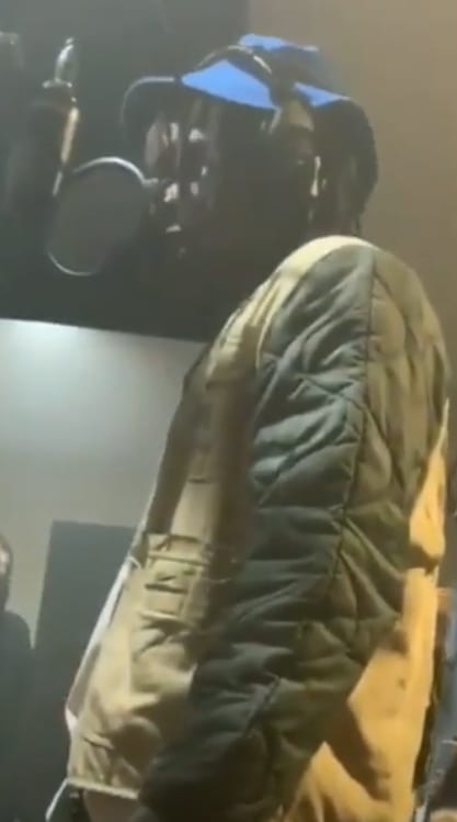 UnoTheActivist stands at a microphone in a recording studio, wearing a blue cap and a quilted tan and black jacket, dimly lit.
