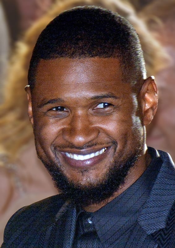 Usher smiles broadly at the camera in a close-up portrait, wearing a dark textured jacket over a black shirt.