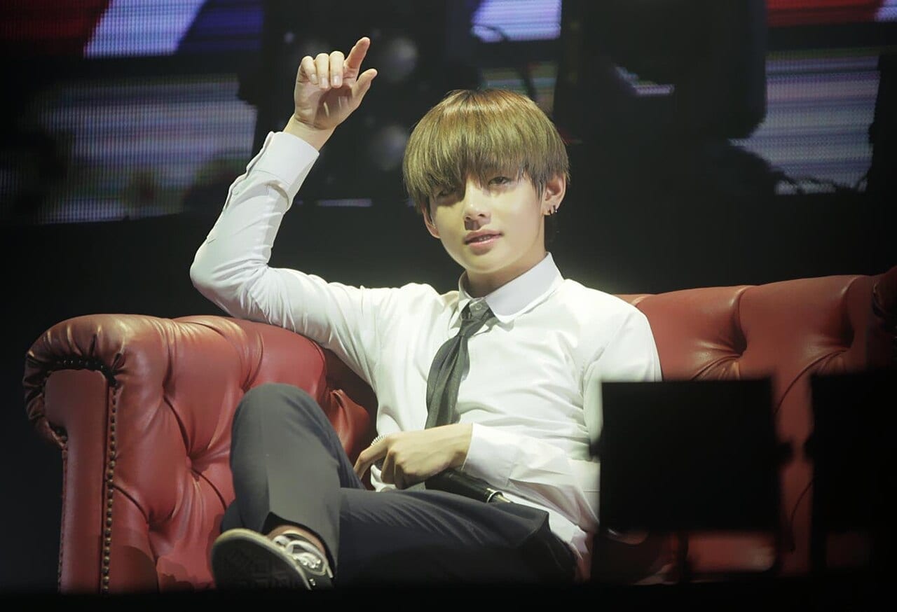 V of BTS sits on a red leather sofa on stage in a white shirt and tie, raising one hand.