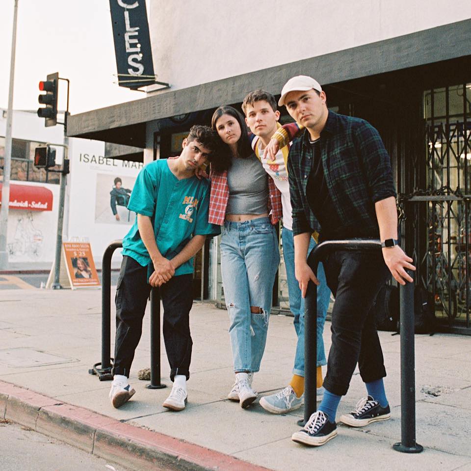 The four members of Valley stand against bike racks on a sunlit sidewalk outside a corner shop, three in flannel and T-shirts and one in a white cap.