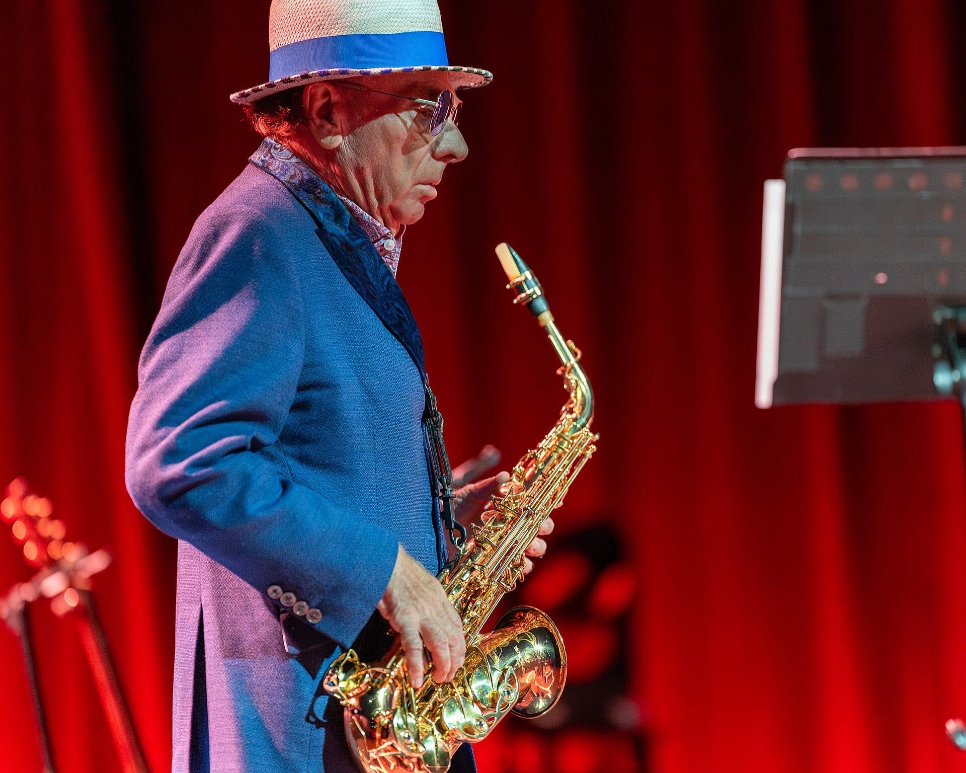Van Morrison plays a gold saxophone in profile, wearing a straw fedora and tinted glasses, lit against a red stage curtain.