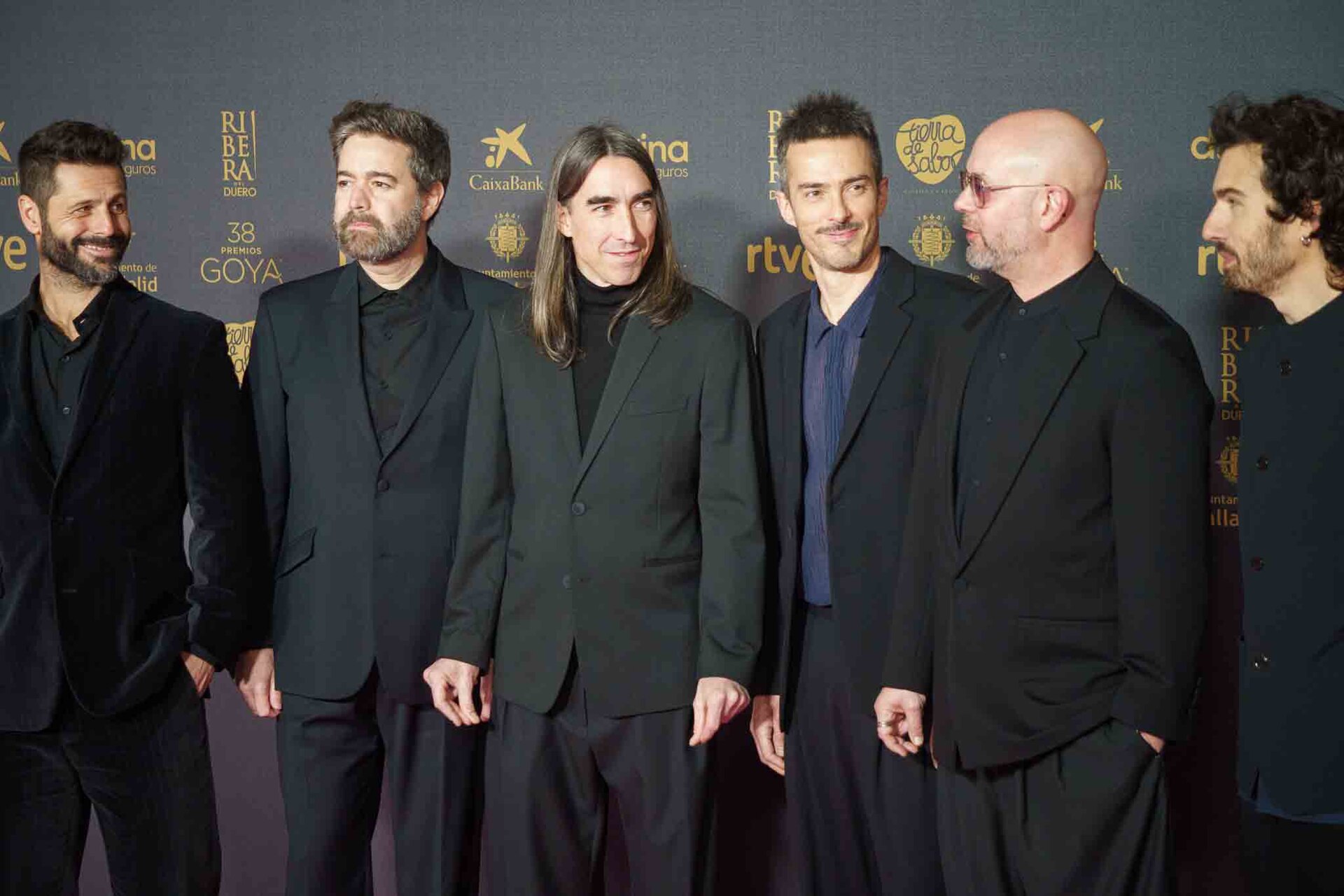 Six men in black suits stand in a line on a red carpet in front of a sponsor backdrop, several glancing sideways at each other