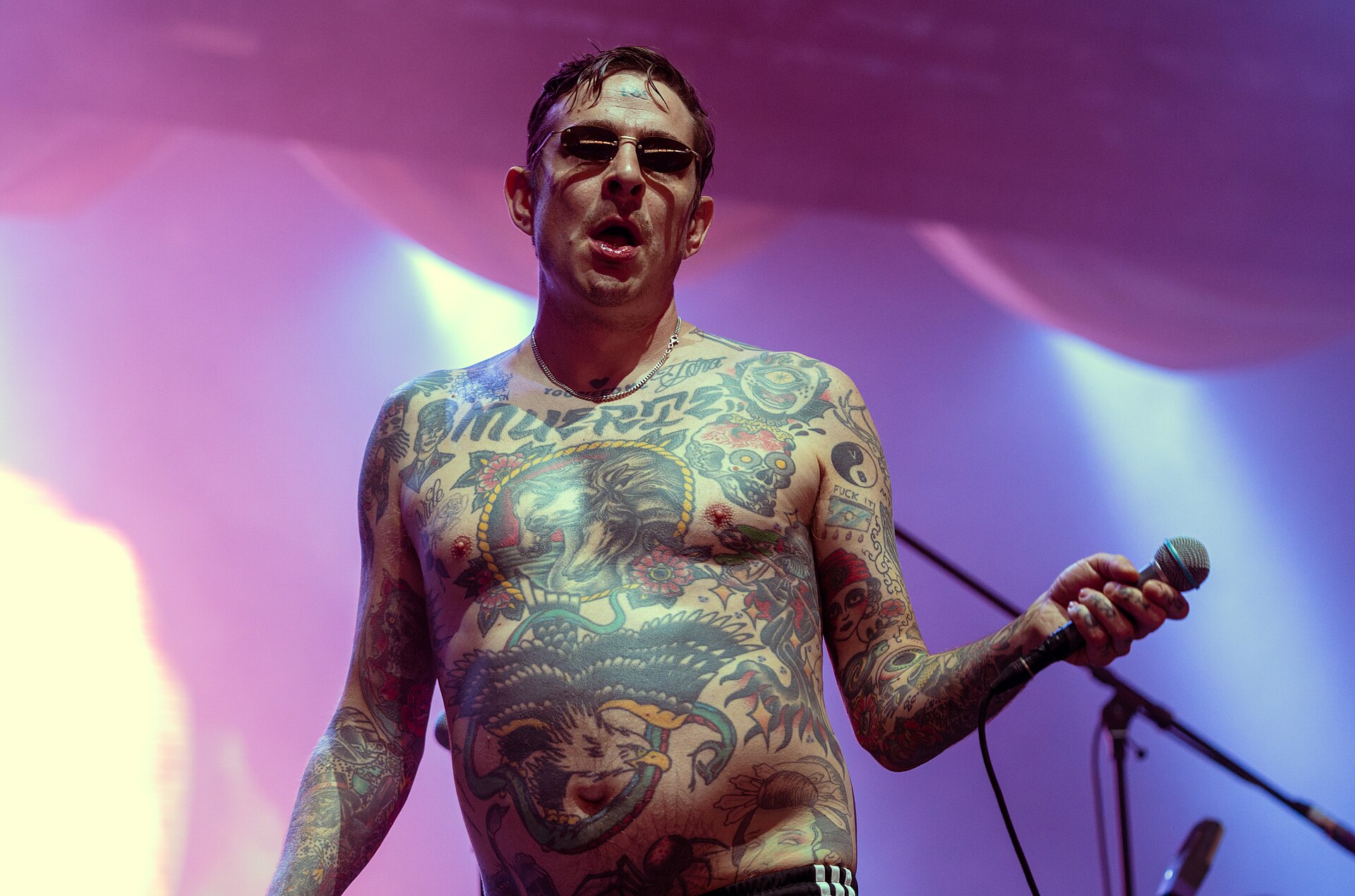 The shirtless, heavily tattooed singer of Viagra Boys stands at a microphone stand in dark sunglasses, mouth open mid-line, against a pink and purple lit backdrop.