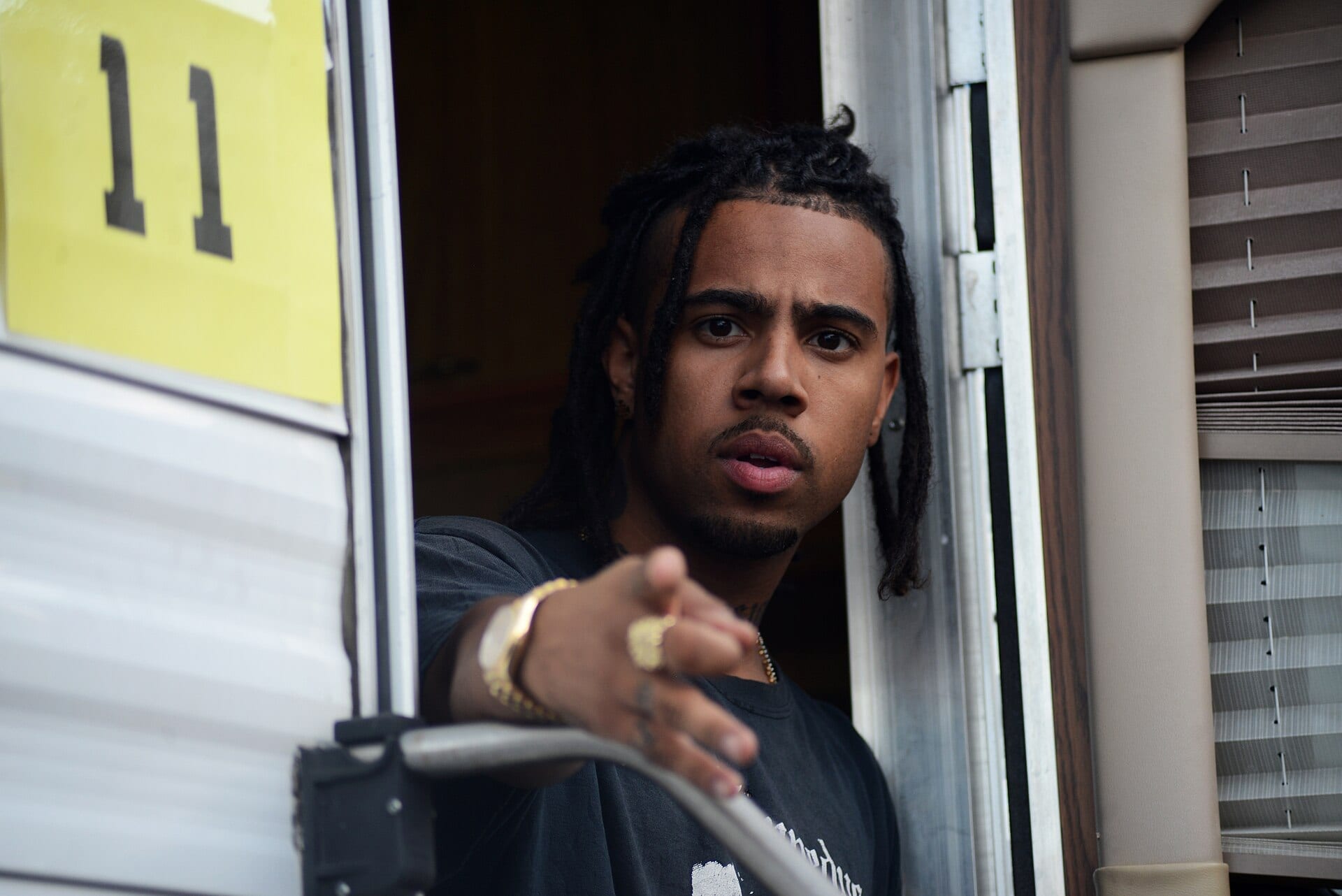 Vic Mensa leans out of a doorway marked '11,' pointing toward the camera, wearing gold jewelry and a black T-shirt.