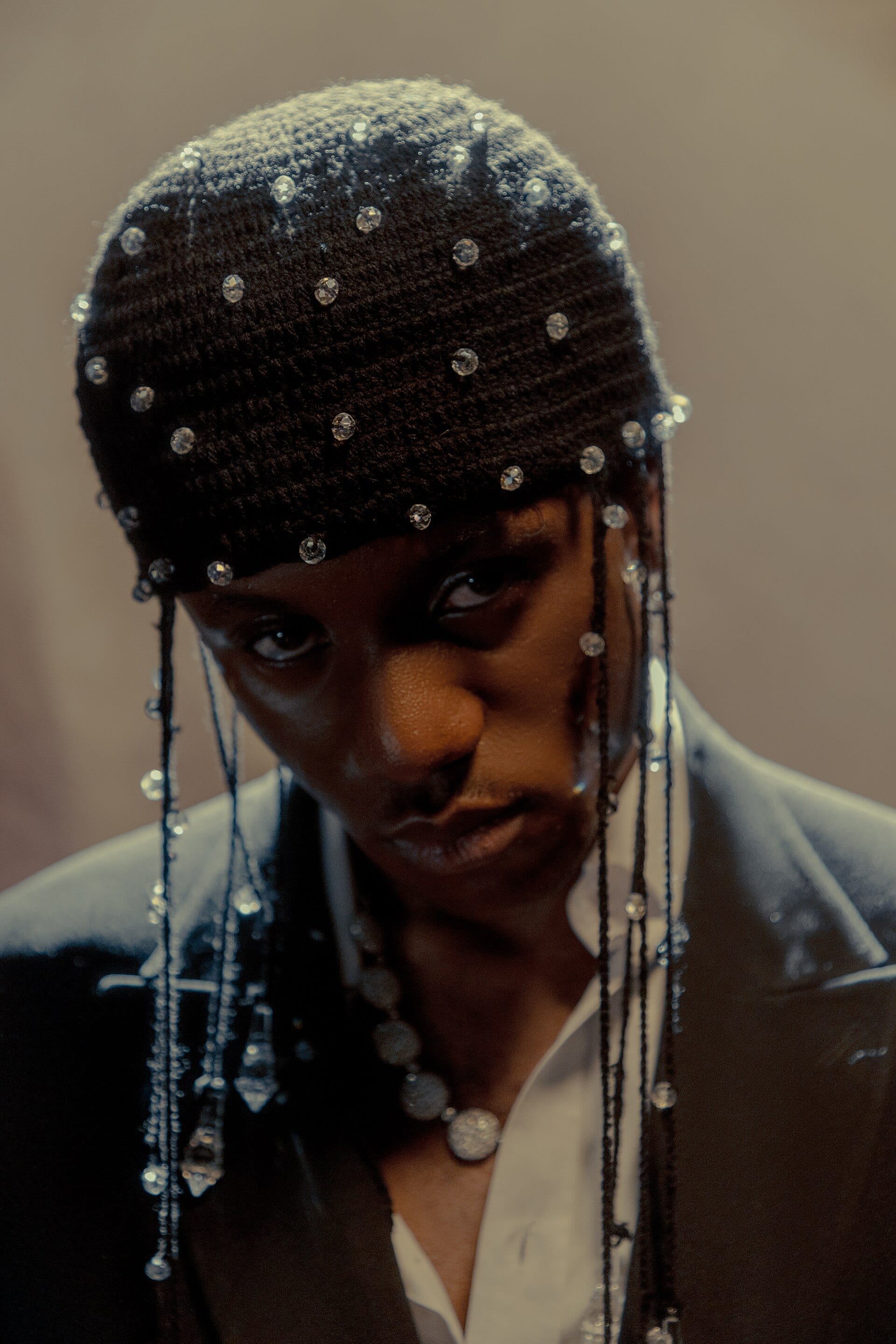 Victony looks into the camera in a beaded black crochet cap, strands of crystals falling past his face over a dark blazer and open white shirt.