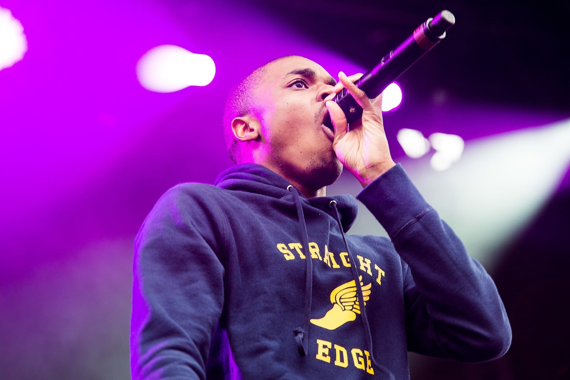 Vince Staples, with a shaved head and wearing a navy "Straight Edge" hoodie, sings into a microphone under purple stage lighting.