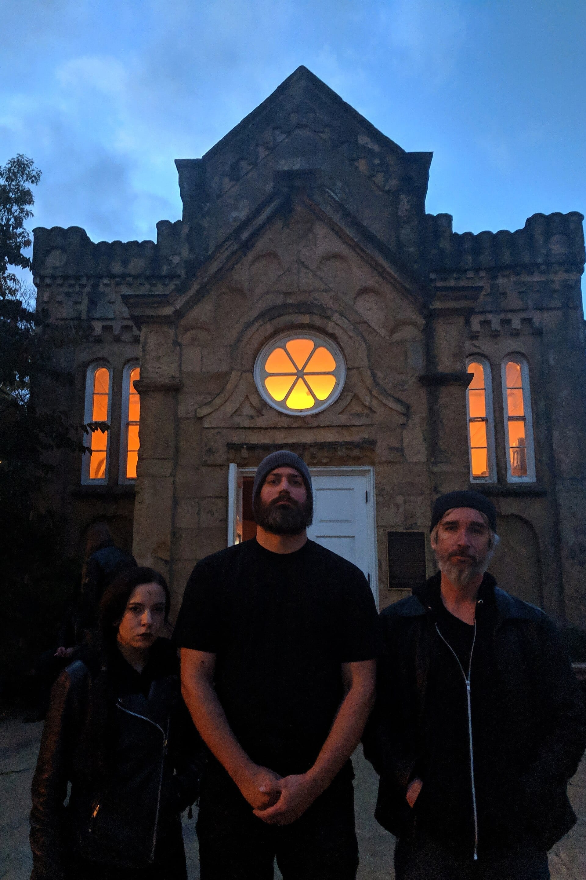 The three members of Volur, dressed in black, stand in front of an old stone building lit orange from within at dusk.