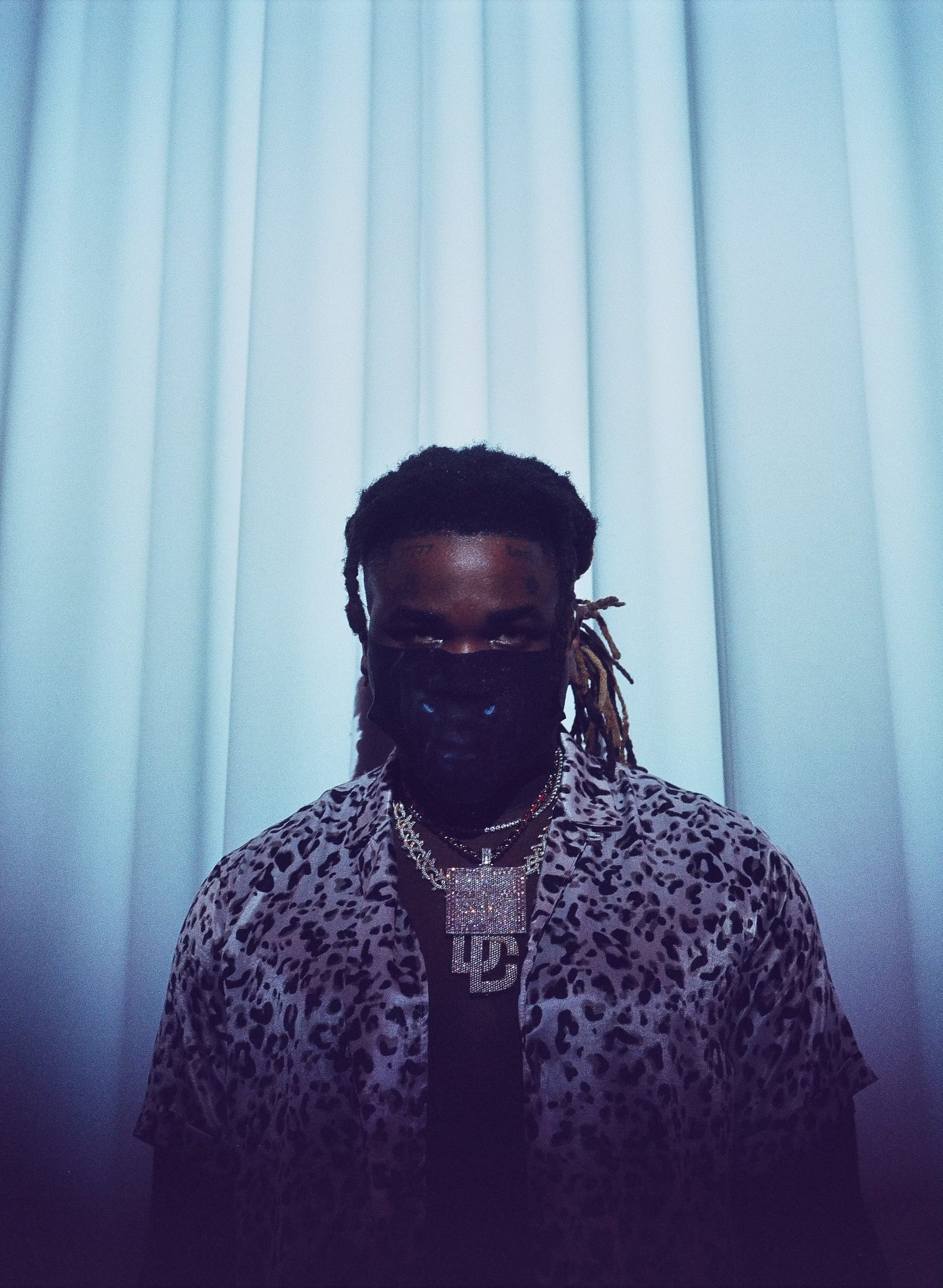 Vory stands against pale blue curtains in a leopard-print shirt and layered chains, the lower half of his face behind a black mask, forehead tattoos and blond-tipped locs framing his eyes.