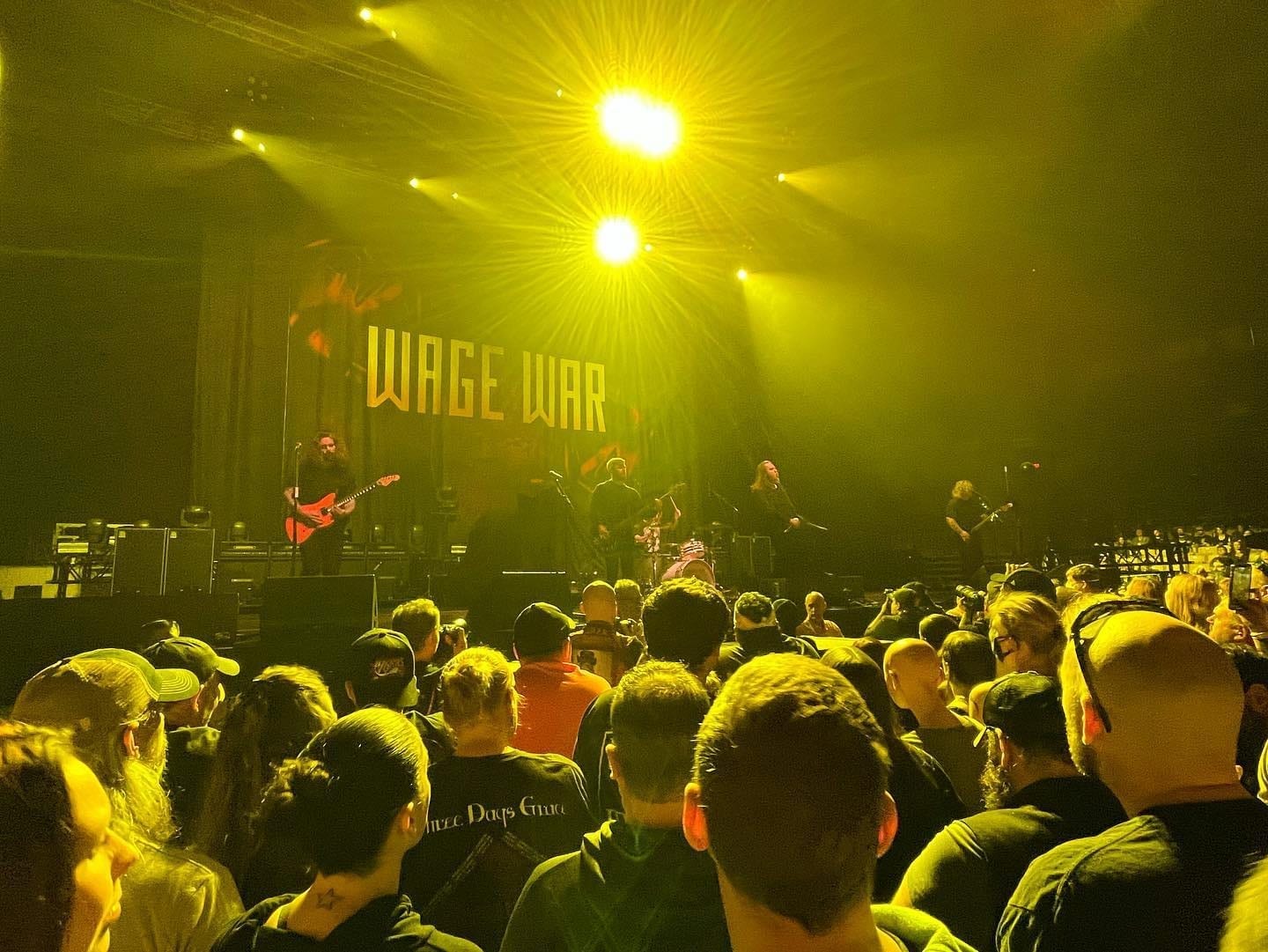 Wage War performs on stage under yellow lights, with a large lit-up WAGE WAR backdrop behind the band and a crowd filling the foreground.