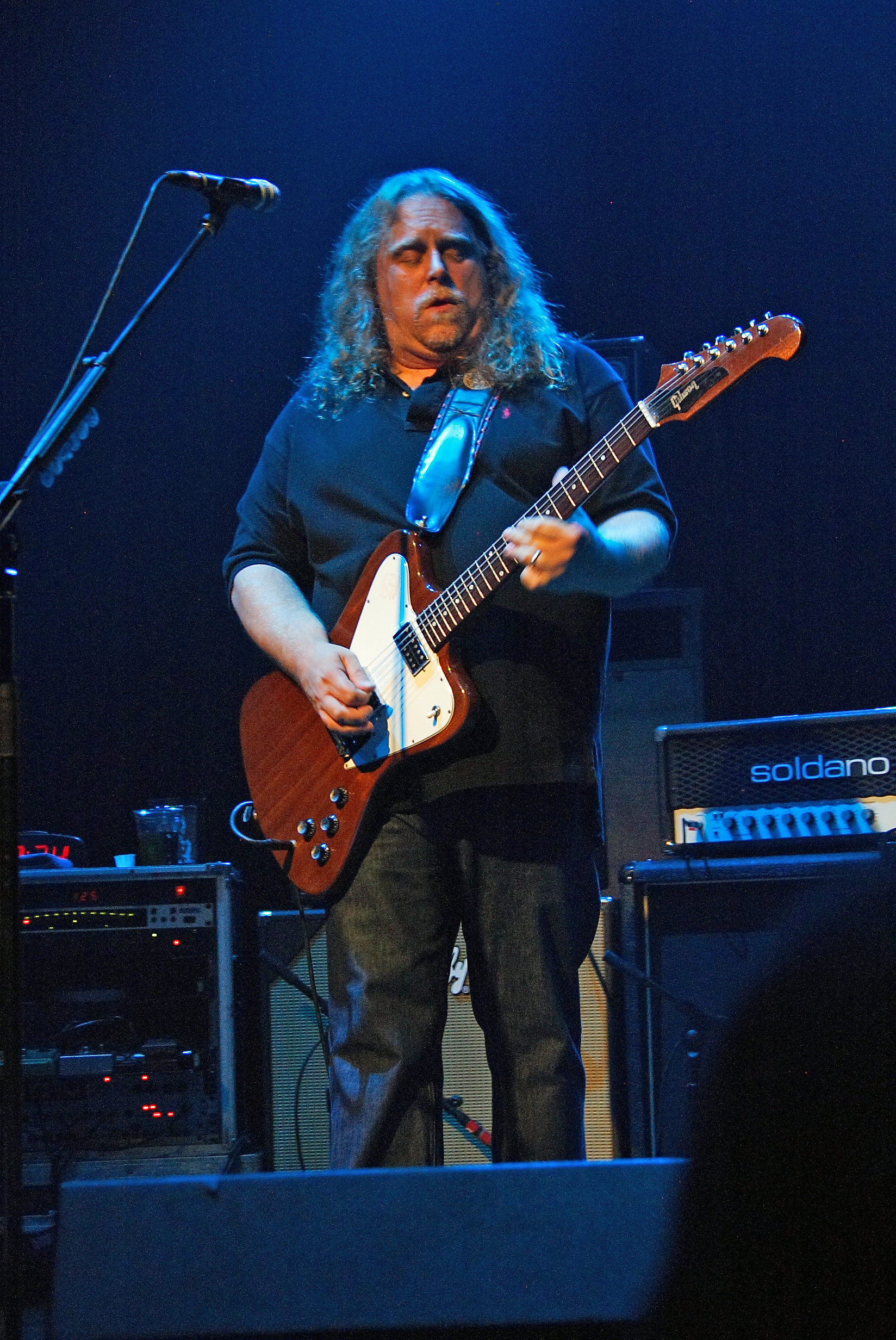 Warren Haynes plays a Gibson Firebird on a blue-lit stage, long curly hair falling forward, a Soldano amp head behind him.