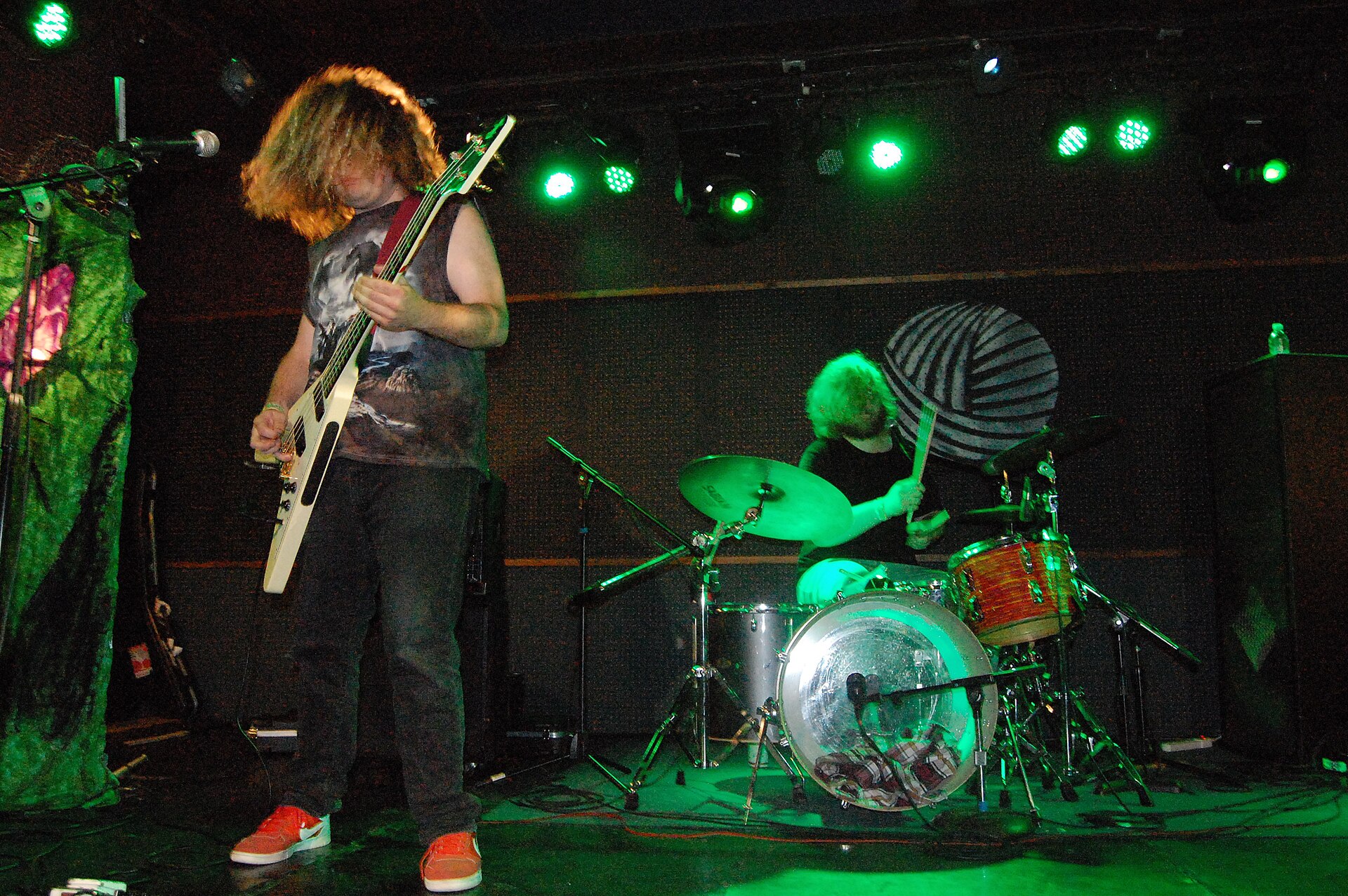 A Wavves guitarist throws his head forward so his hair covers his face, playing a white guitar beside a drummer under green stage light.