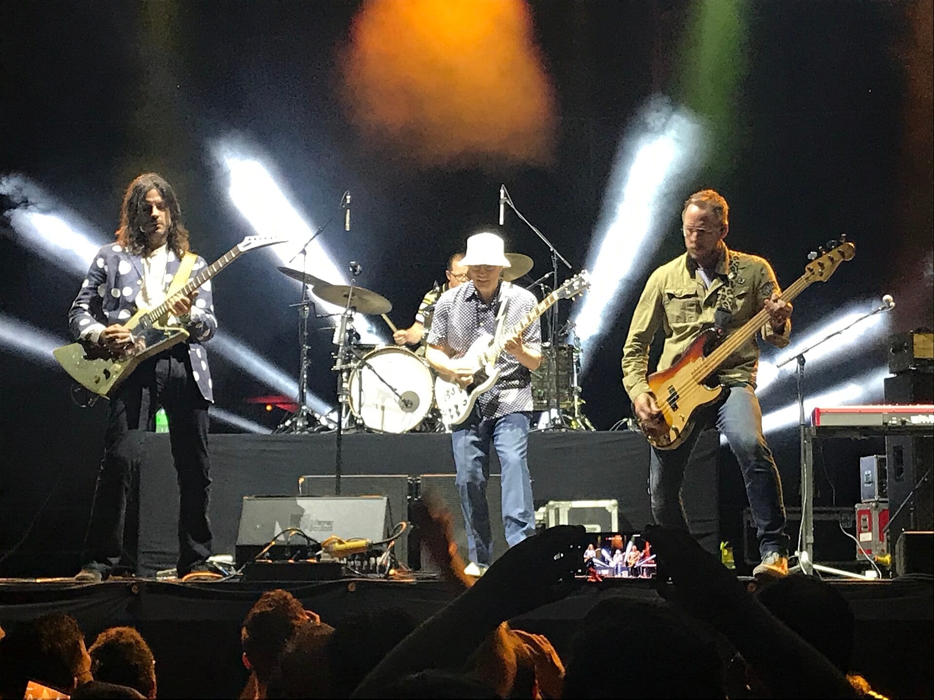 Weezer perform on stage under crossing white stage lights, with Rivers Cuomo in a white hat playing guitar between the bassist and drummer.