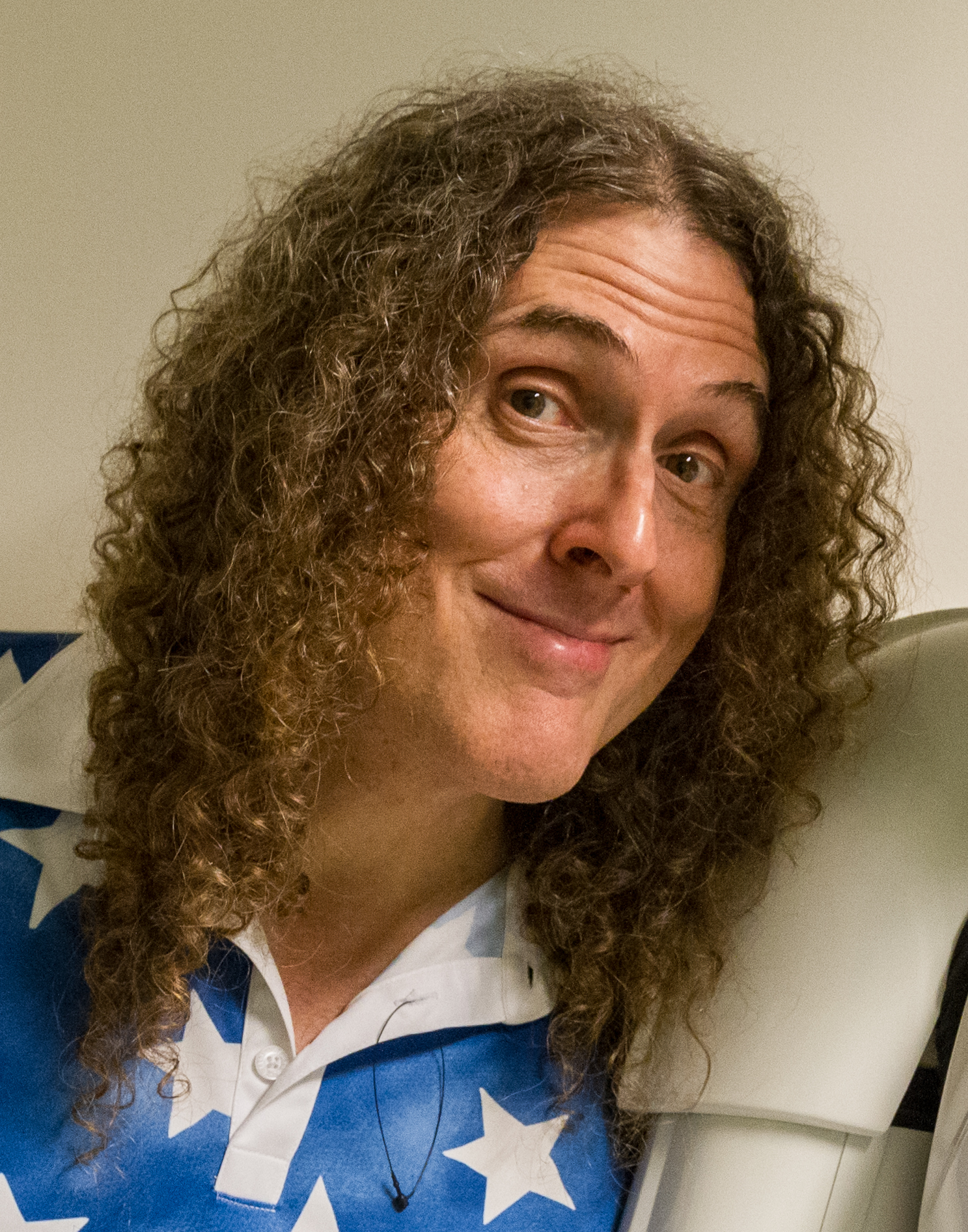 Weird Al Yankovic tilts his head and smiles, long curly hair falling past his shoulders over a blue shirt printed with white stars.
