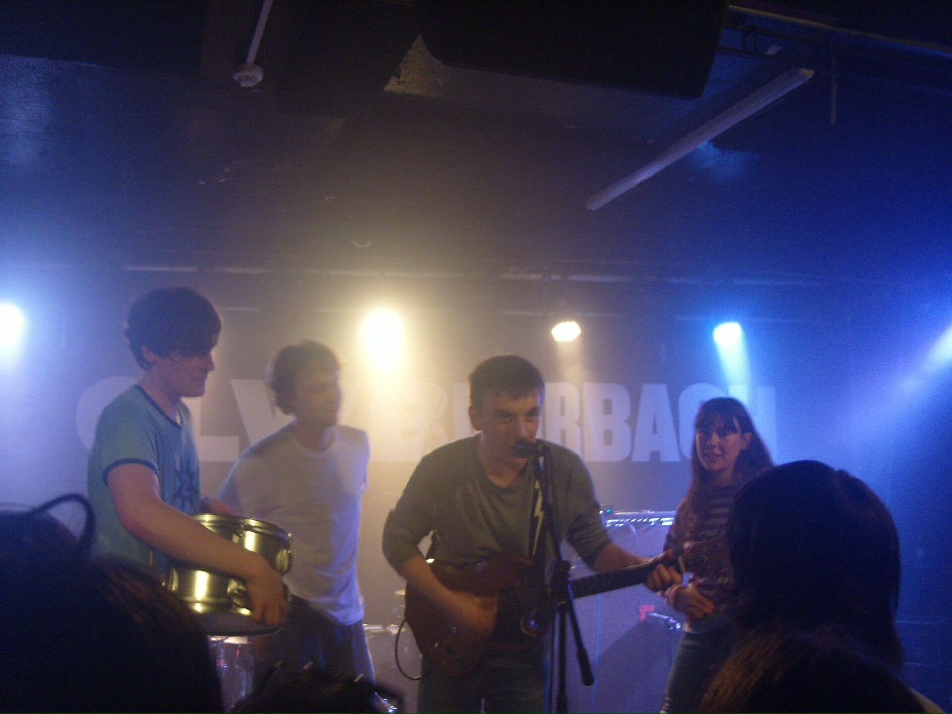 Four members of Westside Cowboy perform on a small stage under blue light: a singer at the mic, a drummer, and two guitarists.