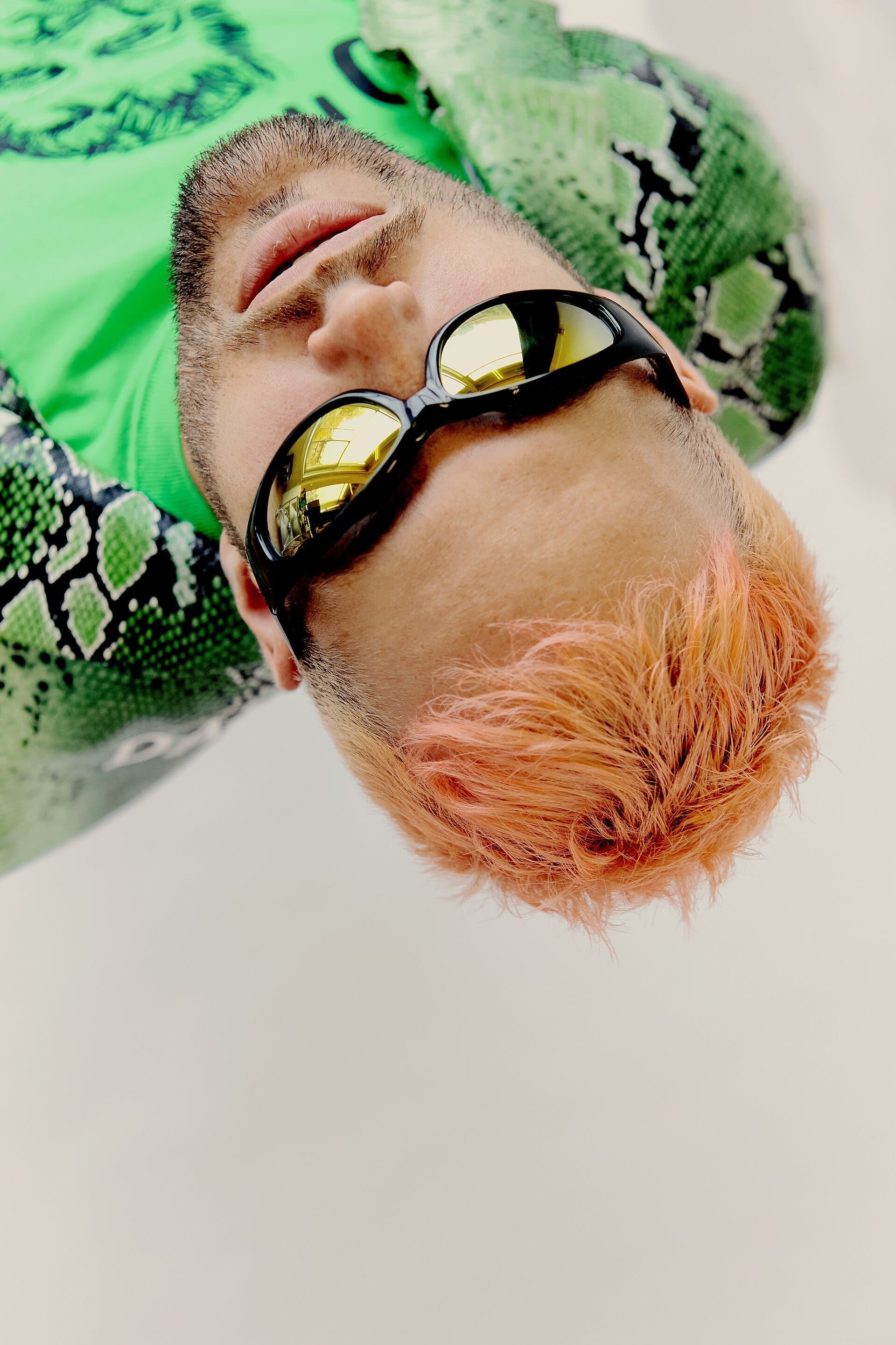 A close-up portrait of What So Not, wearing black sunglasses with yellow lenses, a green snakeskin-print jacket, and a coral-dyed beard.