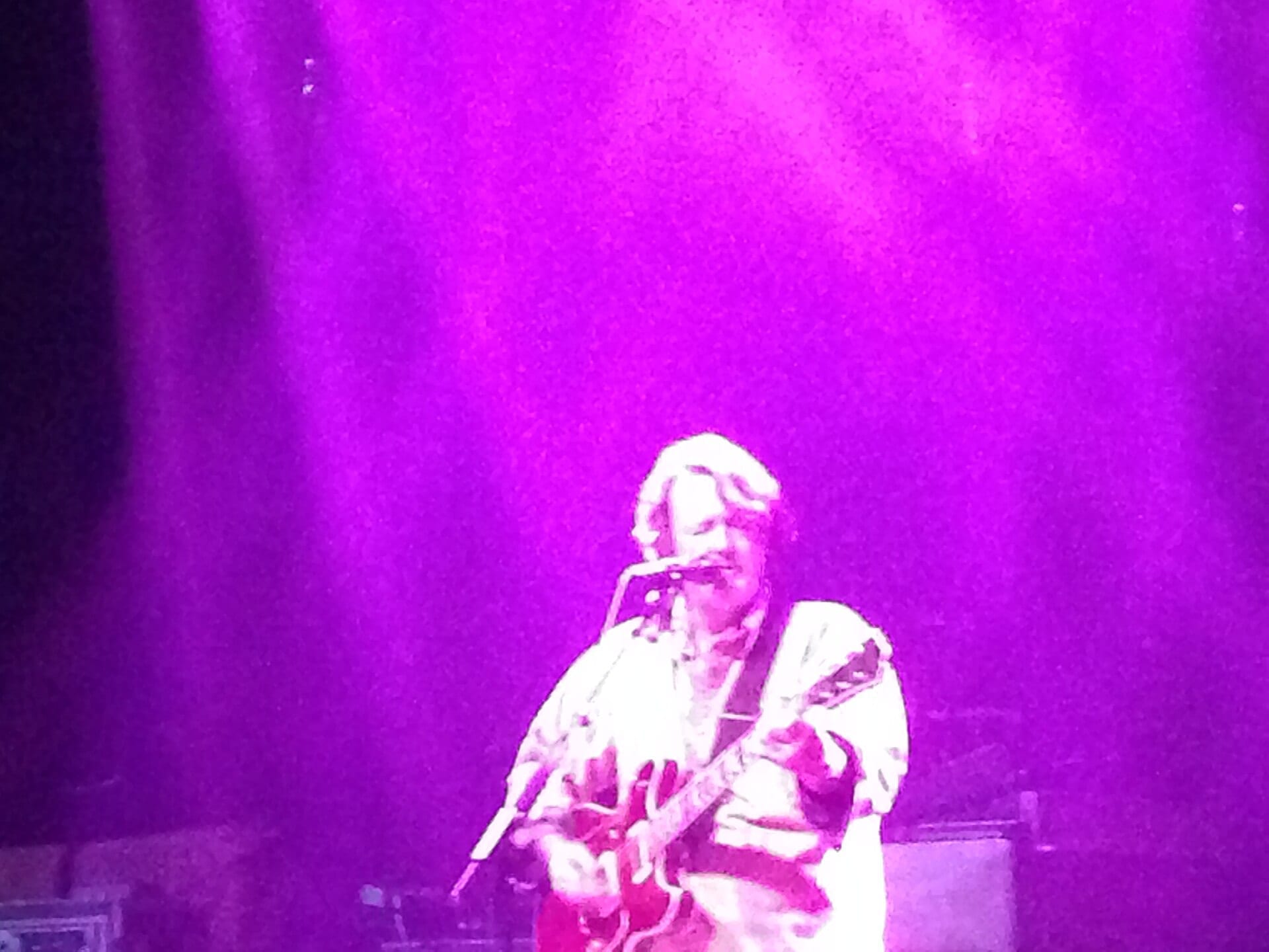John Bell of Widespread Panic sings into a microphone while playing guitar under bright purple stage light.