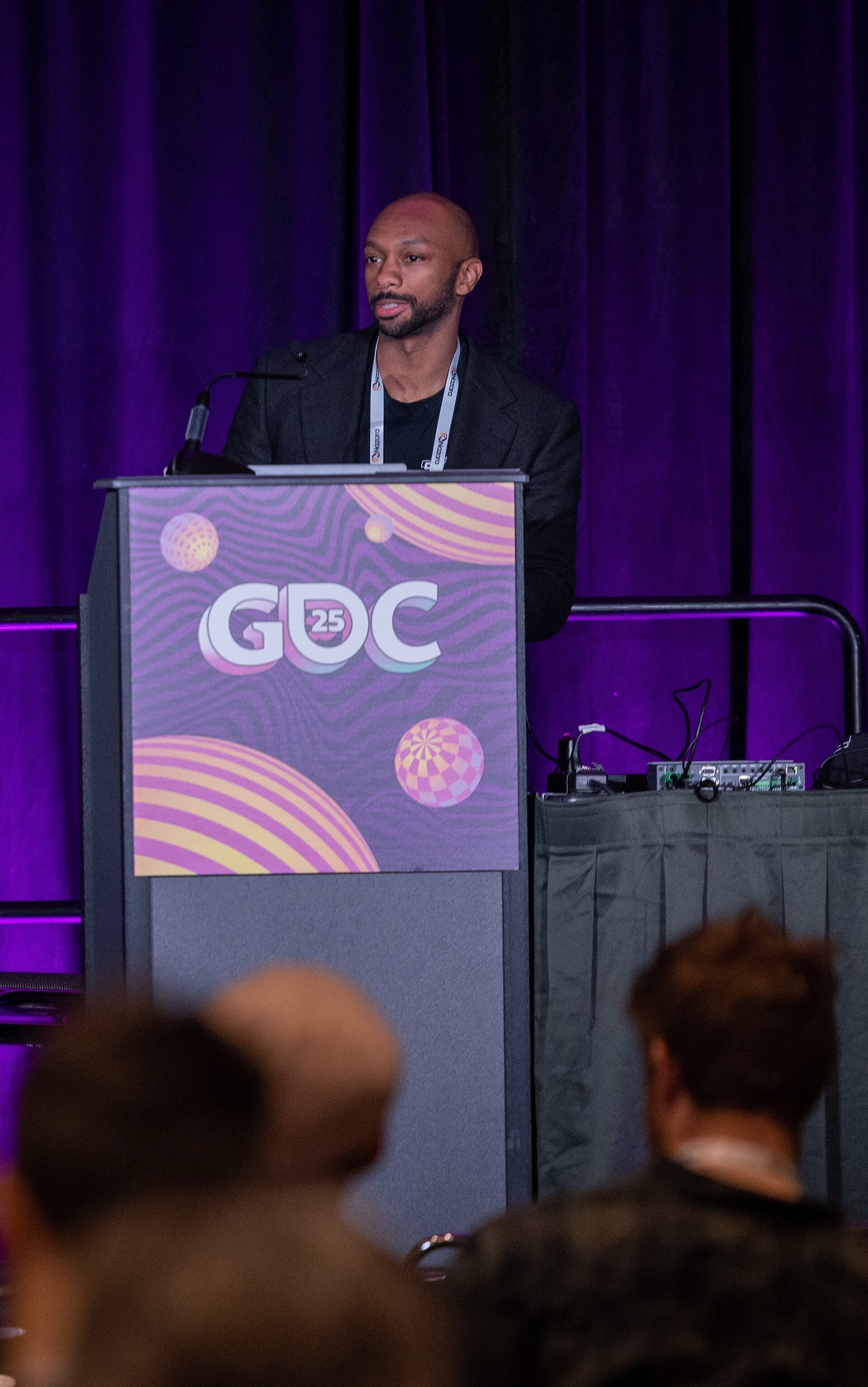 Wilbert Roget II speaks at a lectern branded with the GDC 25 logo, wearing a dark jacket and a conference lanyard.