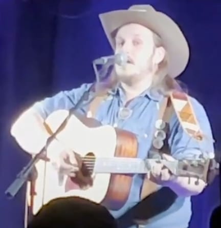 Willi Carlisle sings at a microphone in a pale cowboy hat and a blue snap shirt, strumming an acoustic guitar on a stage lit blue.