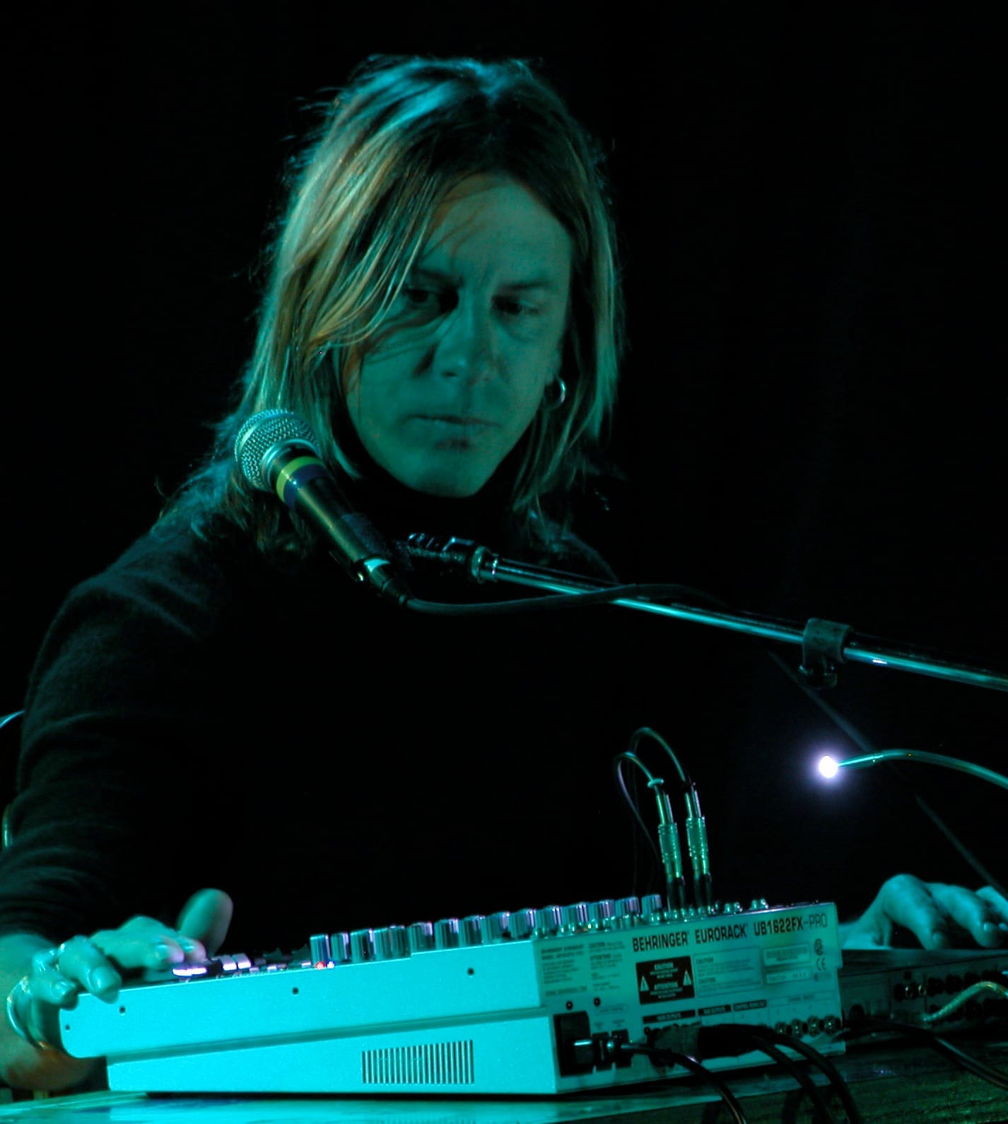 William Basinski works a small mixing desk with both hands in green stage light, long hair falling past his face, a microphone angled toward him.