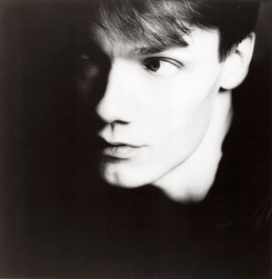 A black-and-white close-up portrait of a young William Orbit, his face lit from one side against a dark background as he looks off to the side.