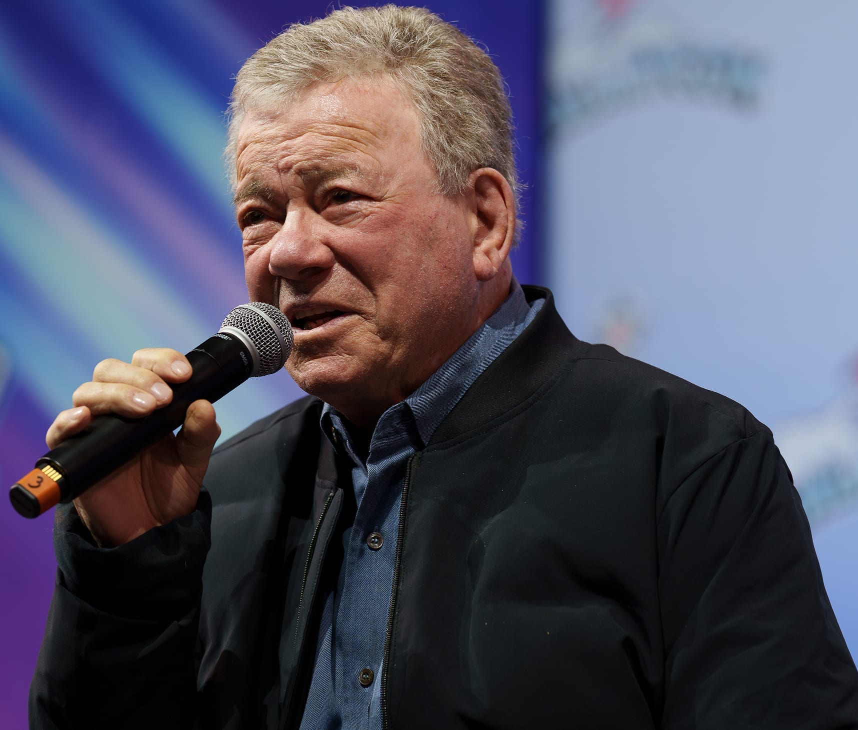 William Shatner speaks into a handheld microphone, wearing a black jacket over a blue collared shirt.