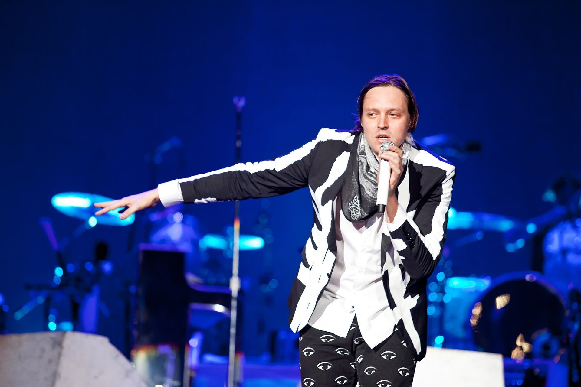 Win Butler sings into a handheld mic with one arm flung out, in a black and white brushstroke-print suit jacket and a grey scarf, blue light behind him.