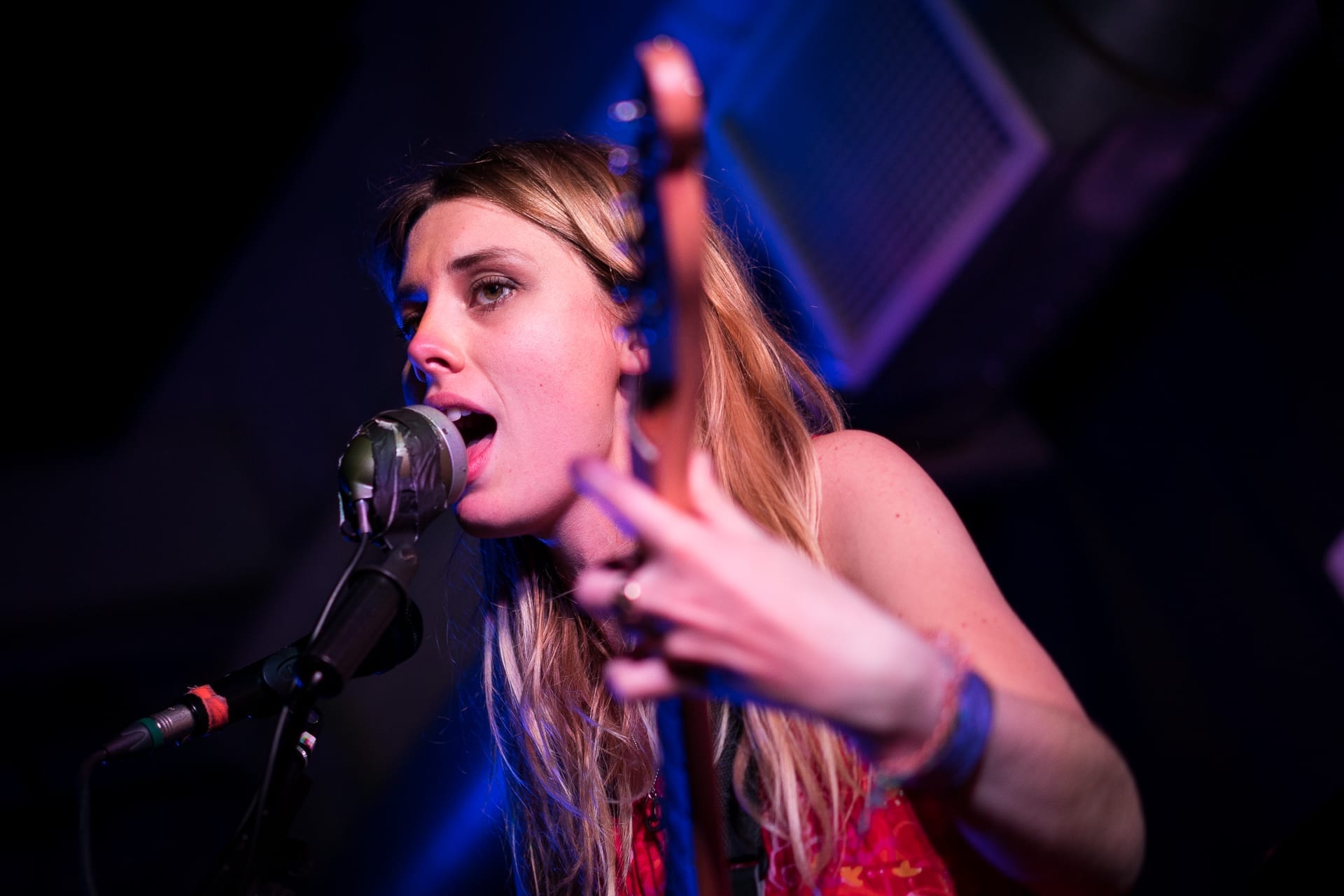 Wolf Alice singer Ellie Rowsell sings close to a microphone while playing guitar under blue and red stage light.