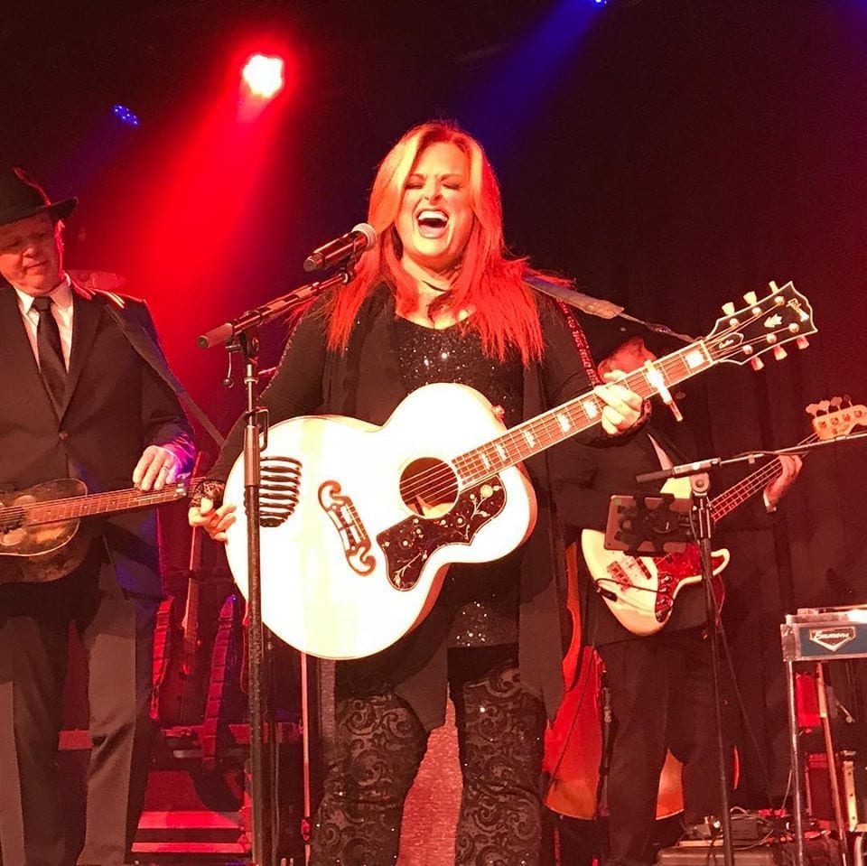 Wynonna Judd sings into a microphone on stage, her red hair lit by stage lighting, with a bandmate playing guitar beside her.