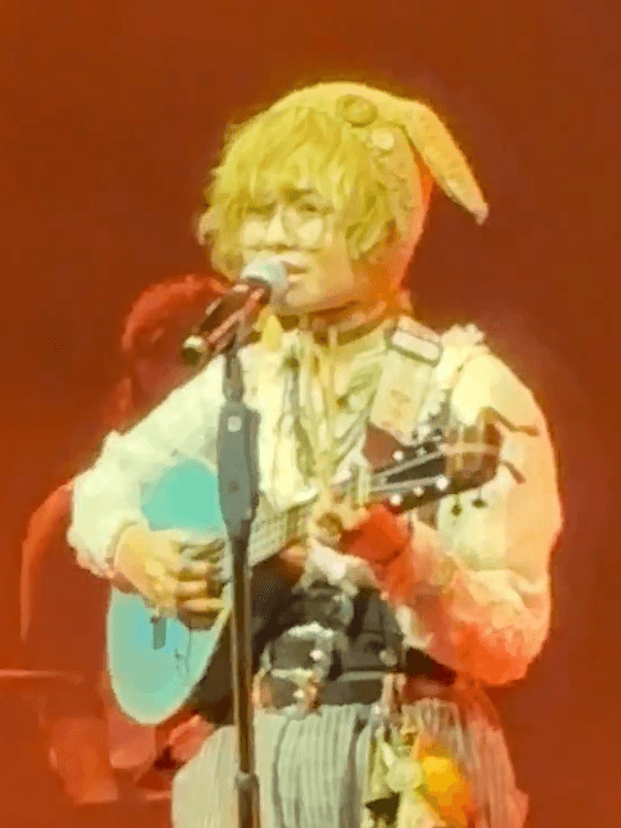 A performer in a rabbit-eared headpiece and ornate costume sings into a microphone while playing a light blue ukulele.