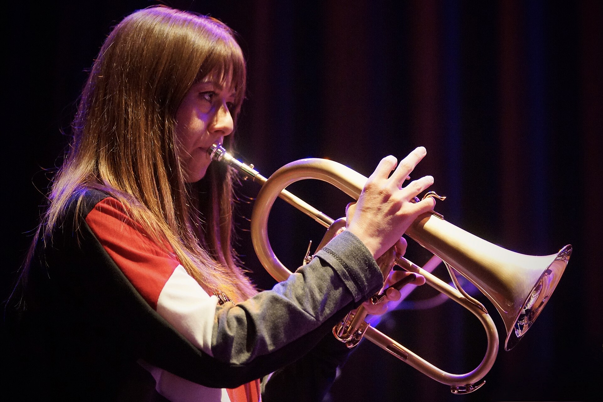 Yazz Ahmed plays a flugelhorn in profile, long hair falling forward, in a black jacket with a red and white stripe.