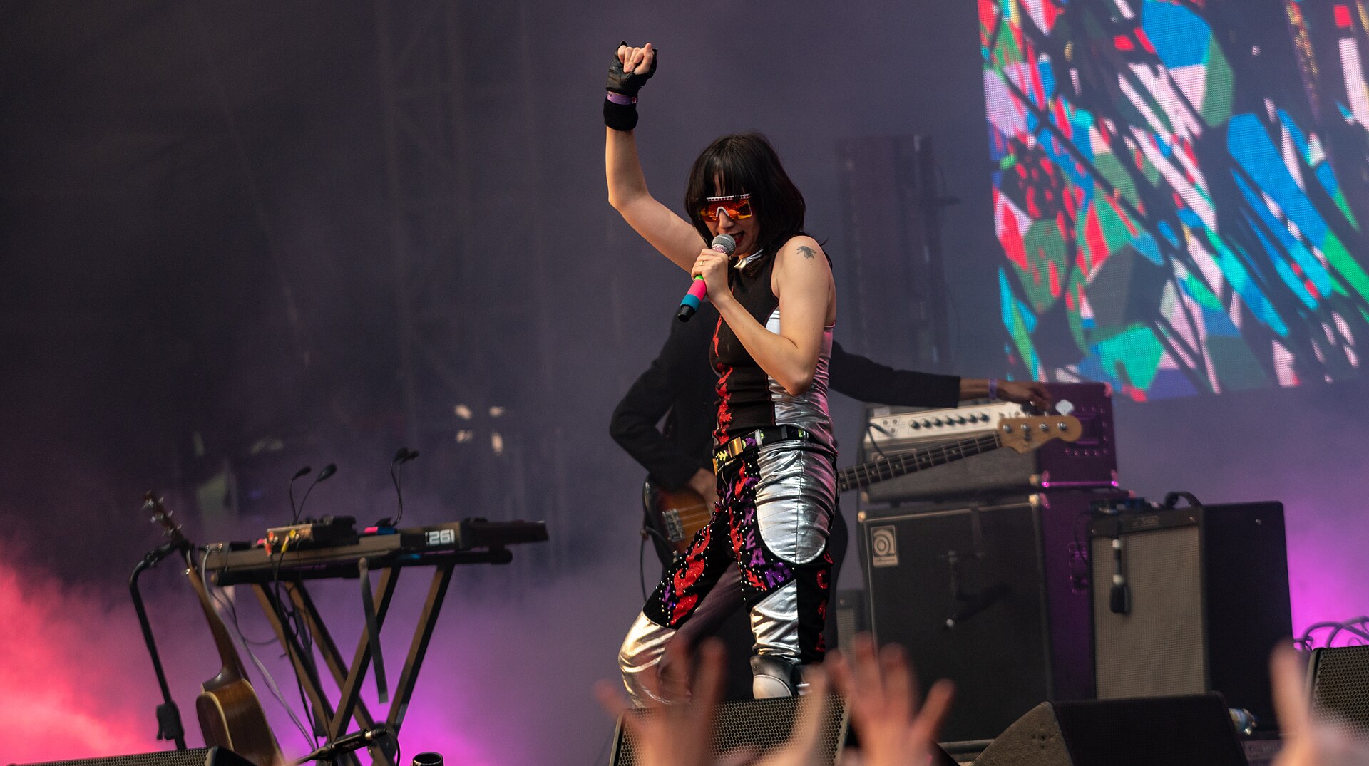 Karen O sings into a microphone with one fist in the air, in visored sunglasses and silver trousers, a keyboard rig beside her.