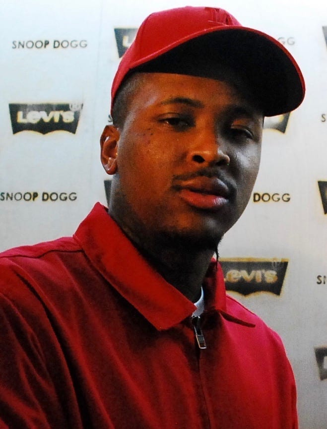 YG in a red cap and red jacket looks toward the camera in front of a step-and-repeat backdrop printed with Snoop Dogg and Levi's logos.