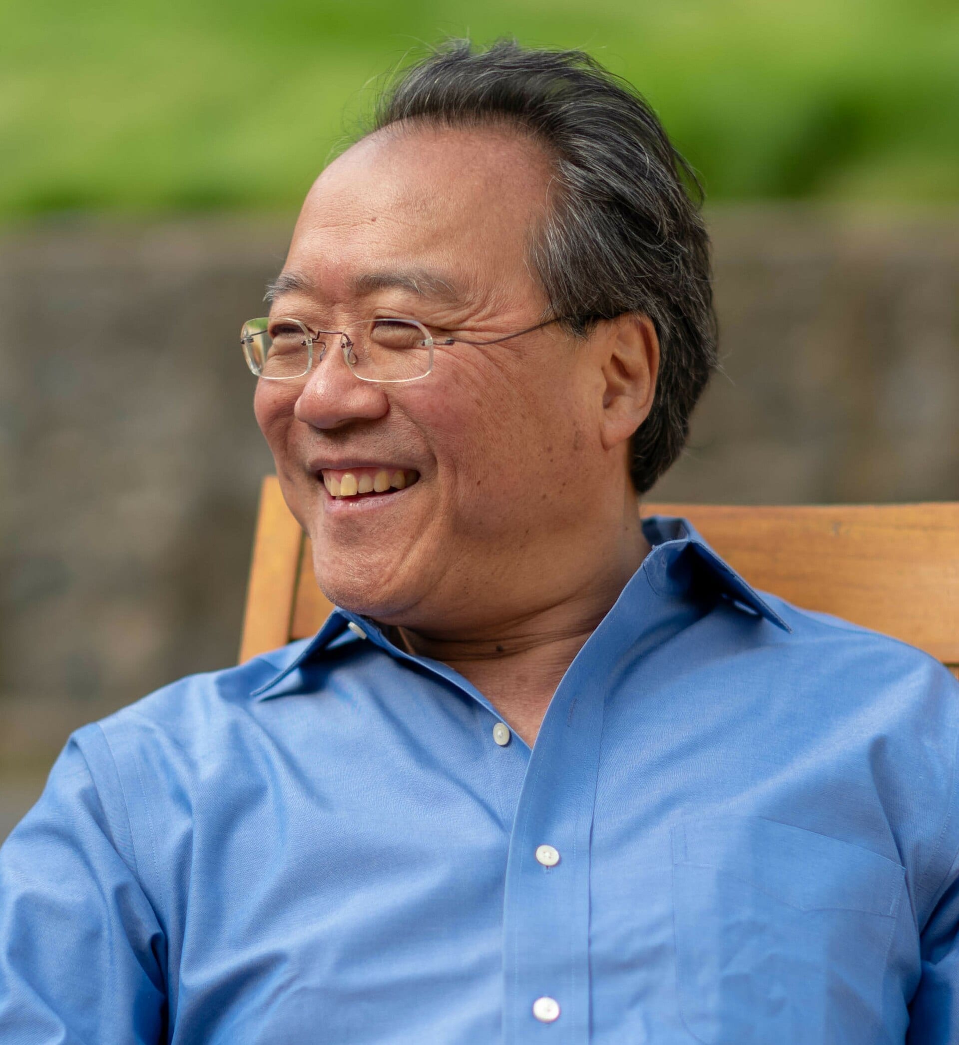 Yo-Yo Ma smiles while seated outdoors, wearing glasses and a blue button-up shirt.