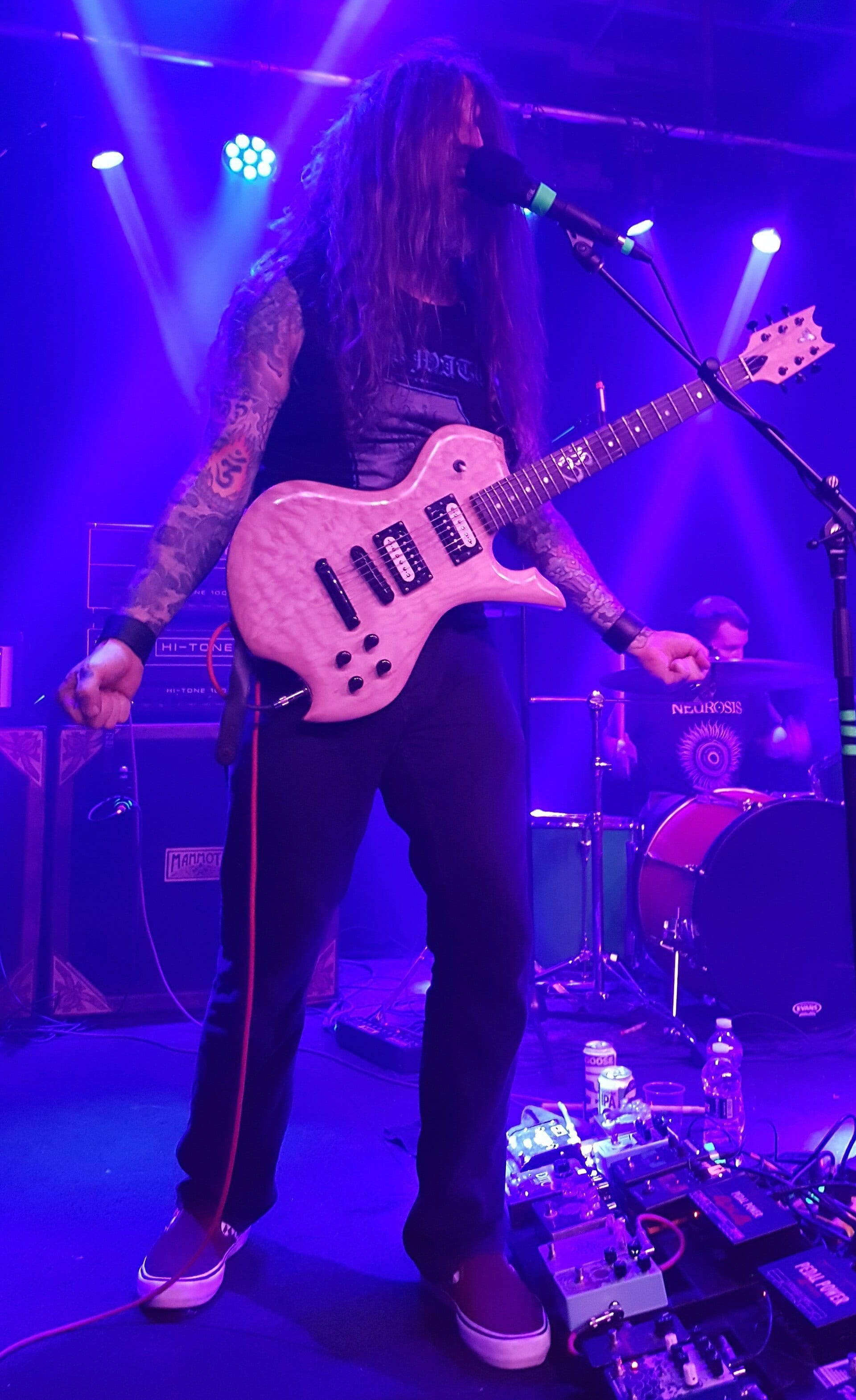 YOB frontman Mike Scheidt plays a light-colored guitar and sings into a microphone under purple stage lighting, his tattooed arms visible.