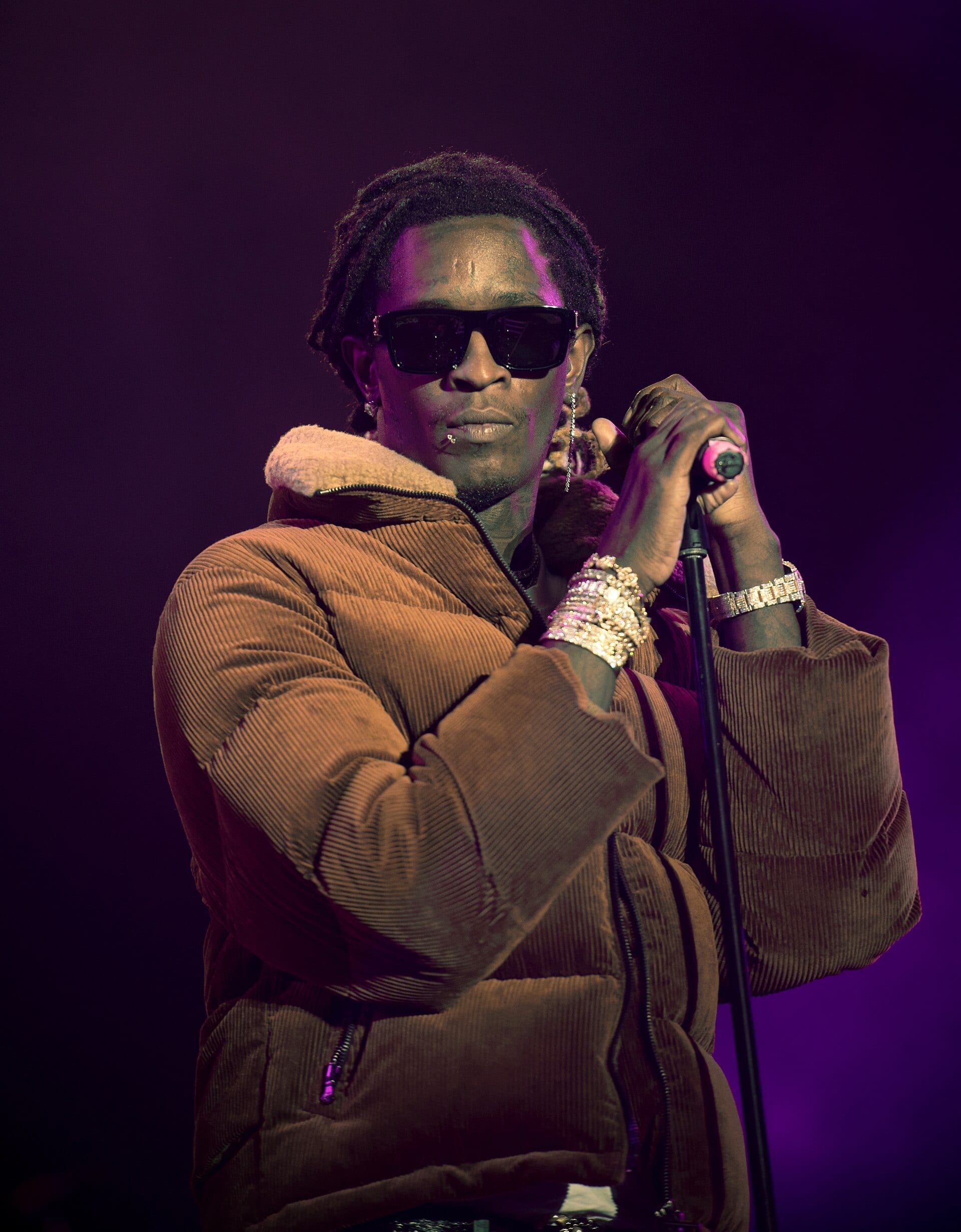 Young Thug, wearing sunglasses and a brown corduroy jacket, holds a microphone stand on stage under purple lighting.