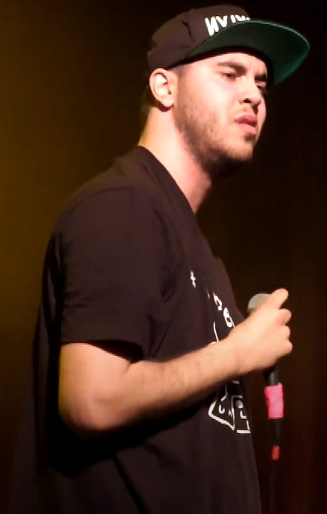 Your Old Droog performs on stage holding a microphone, wearing a dark baseball cap and black t-shirt, photographed from the side.