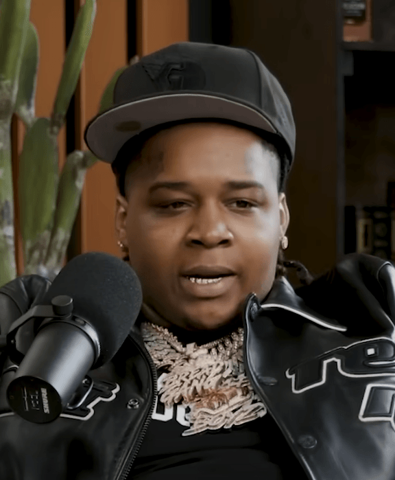 YTB Fatt speaks into a podcast microphone during an interview, wearing a black cap, a black leather jacket and layered chains.