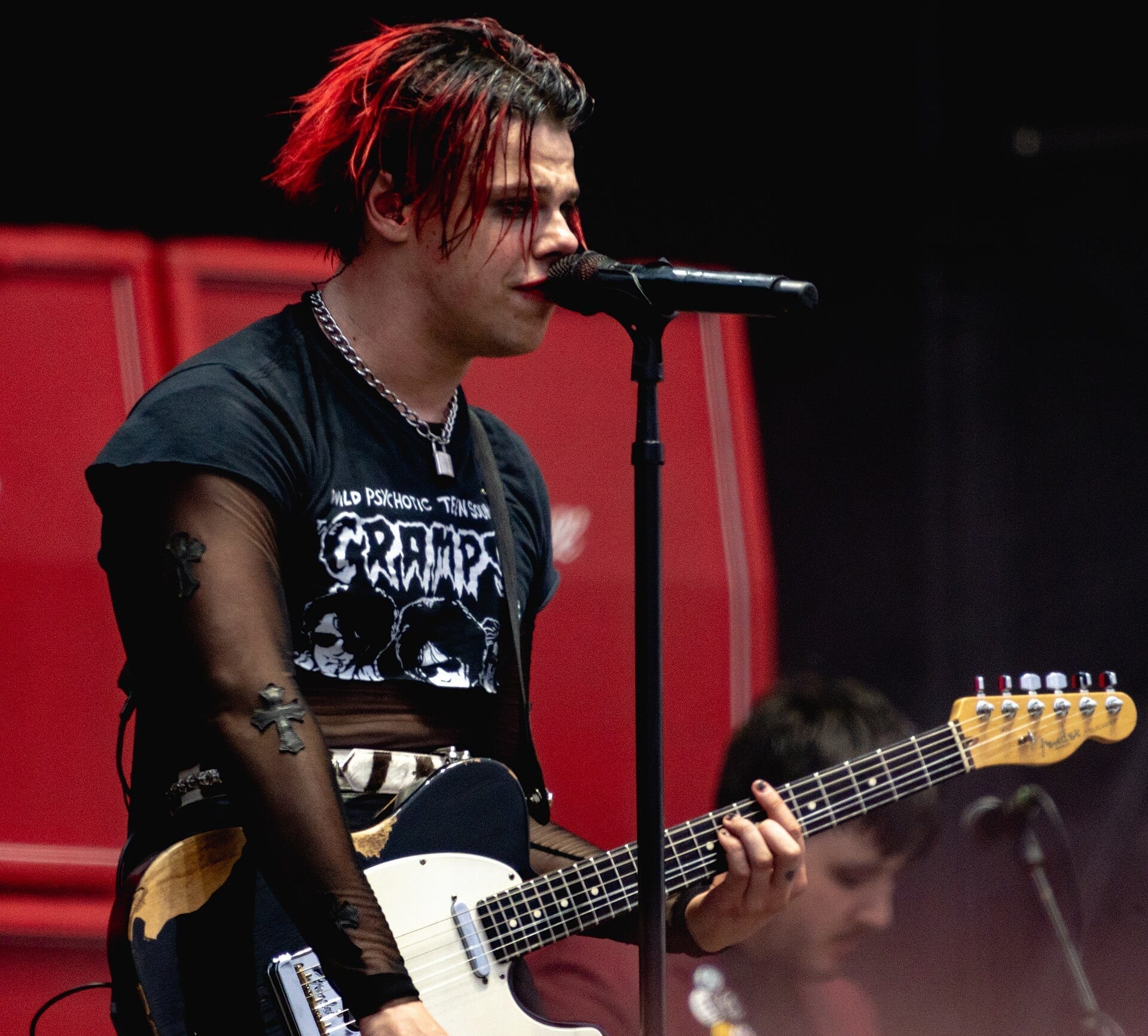 Yungblud, with red-streaked dark hair and a Cramps t-shirt under a sheer black layer, sings into a microphone while playing a white electric guitar.