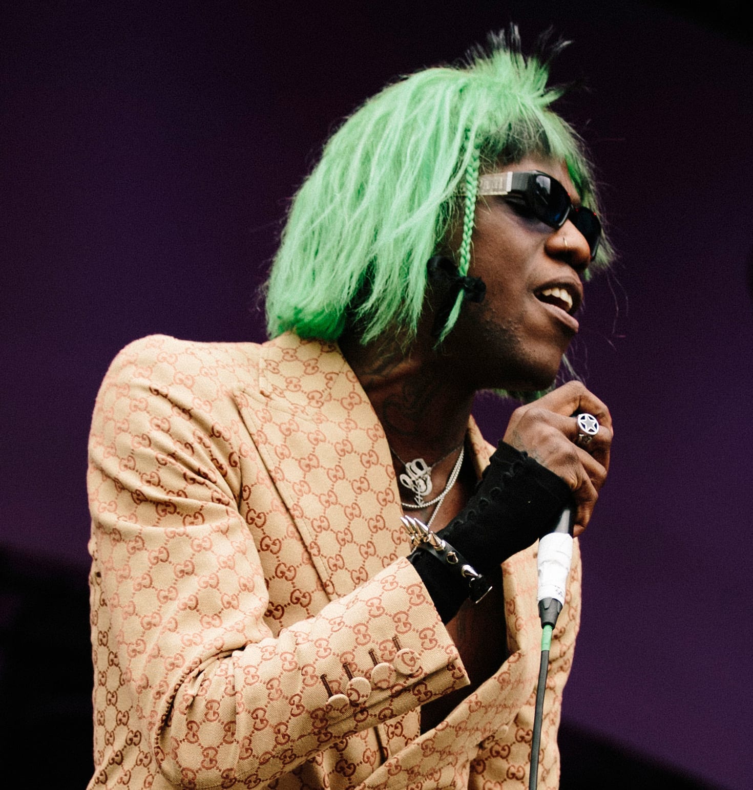Yves Tumor sings into a microphone wearing a patterned tan jacket, dark sunglasses and bright green hair.