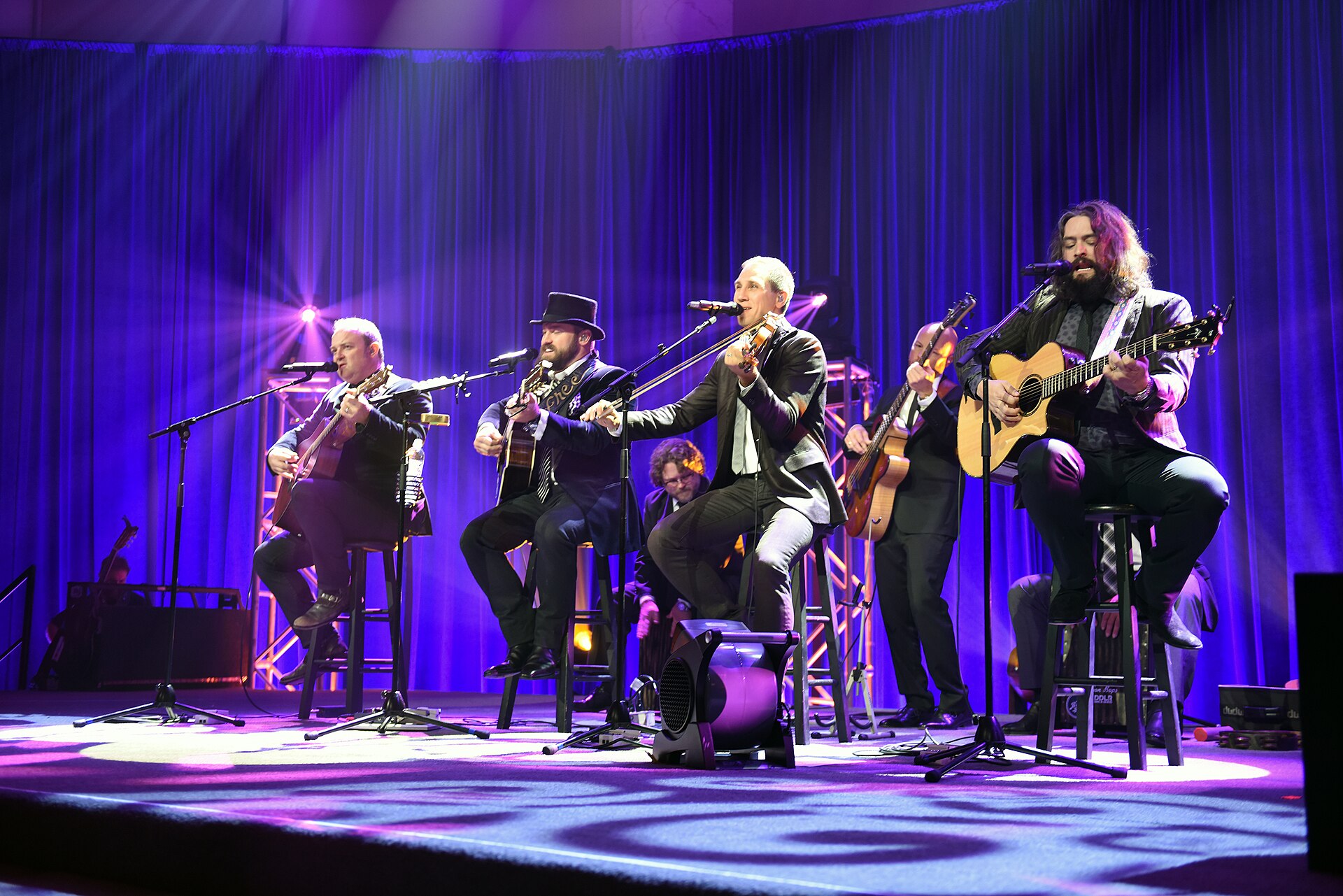 Six members of the Zac Brown Band play a seated acoustic set on stools under purple stage light, Zac Brown in a top hat second from left and a fiddle player centre.