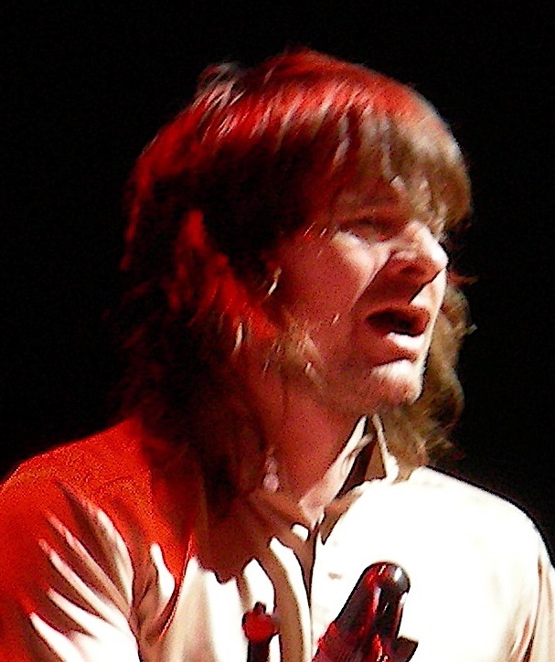 Zak Starkey mid-song with his mouth open and long hair falling across a white shirt, lit red against a black background.