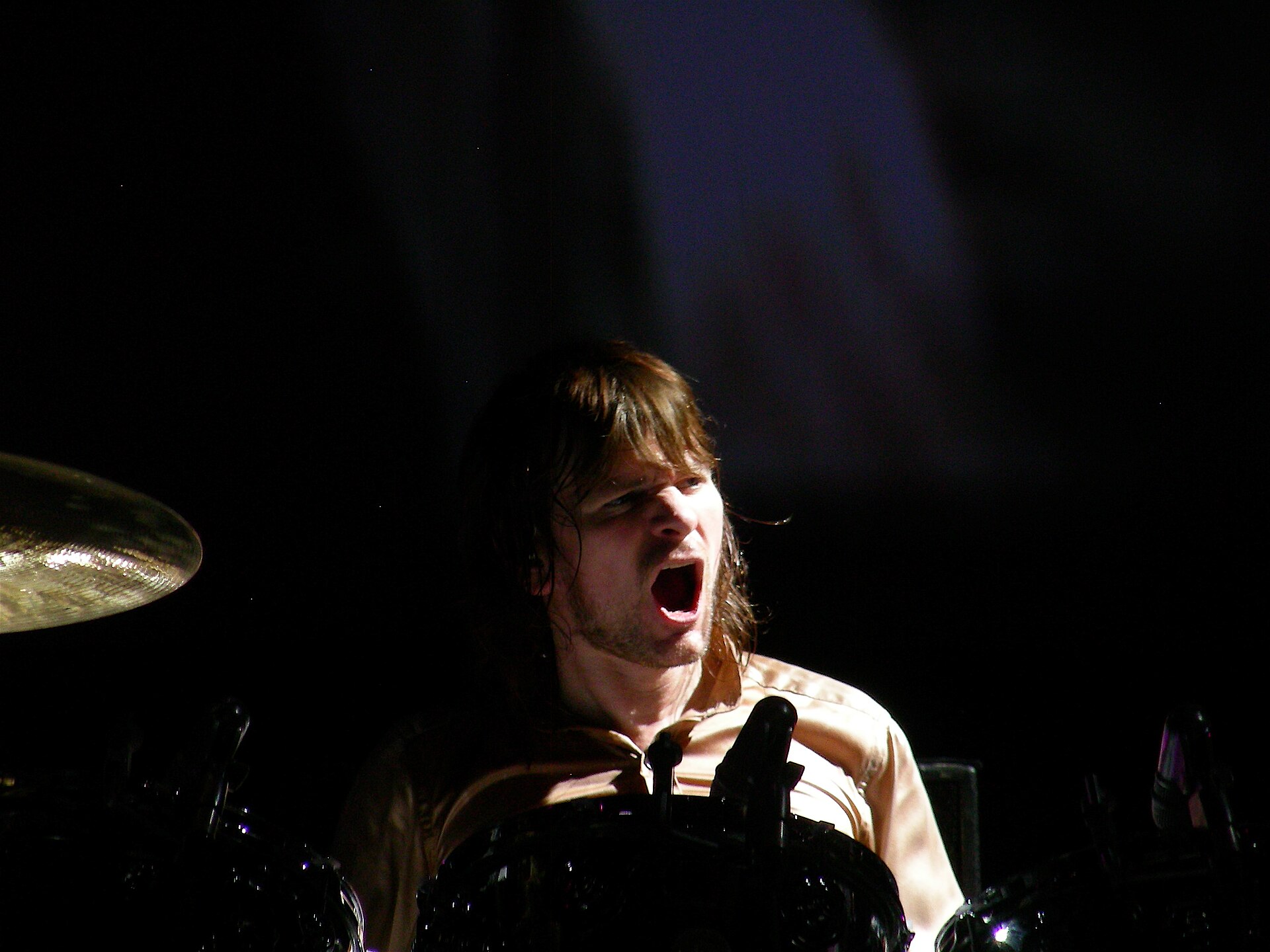 Zak Starkey shouts mid-fill behind a drum kit, long hair swinging, sticks raised, in a dark sleeveless shirt.