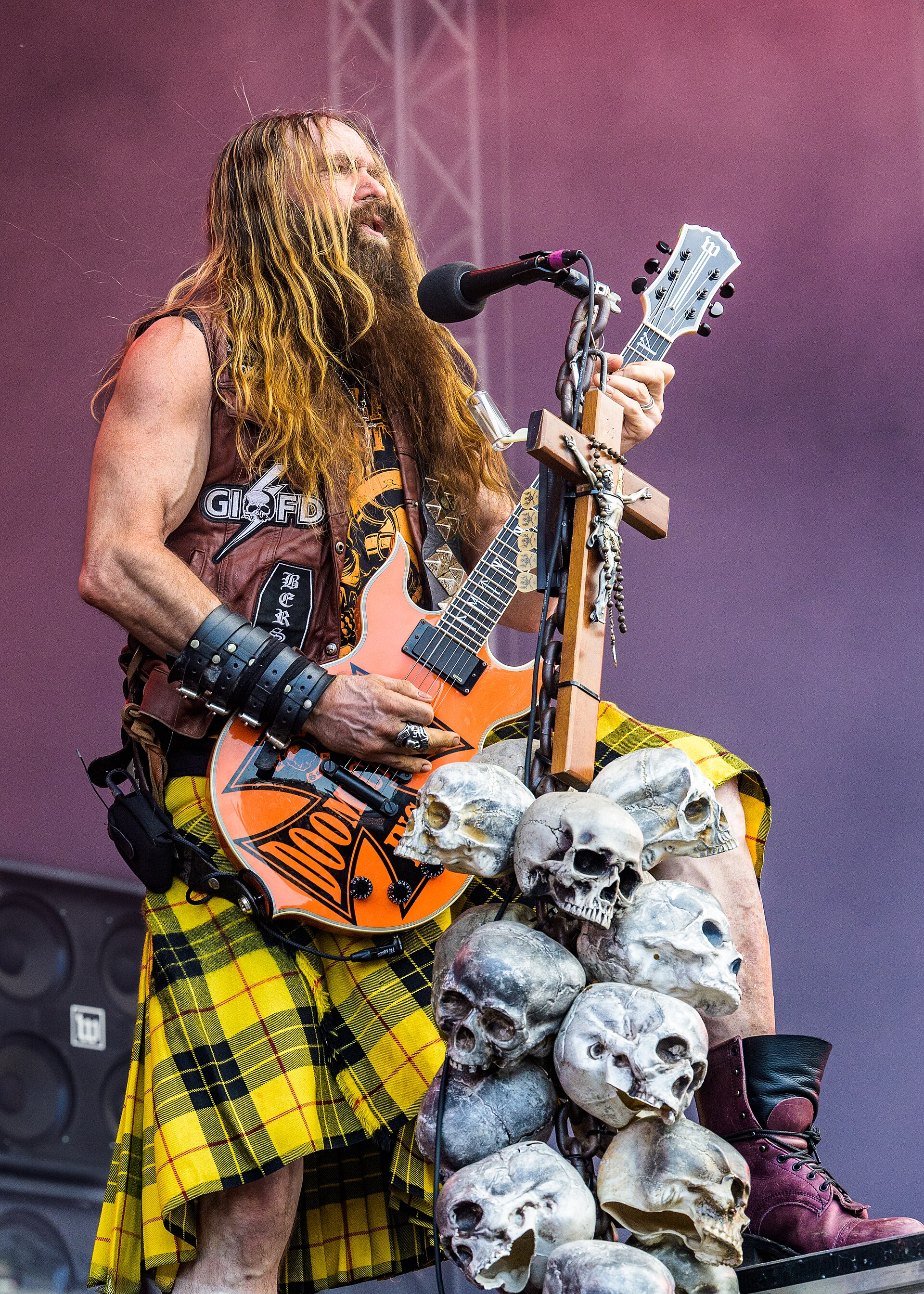 Zakk Wylde singing at the microphone in a yellow tartan kilt and cut-off leather vest, playing an orange bullseye guitar beside a mic stand hung with a crucifix and a chain of skulls