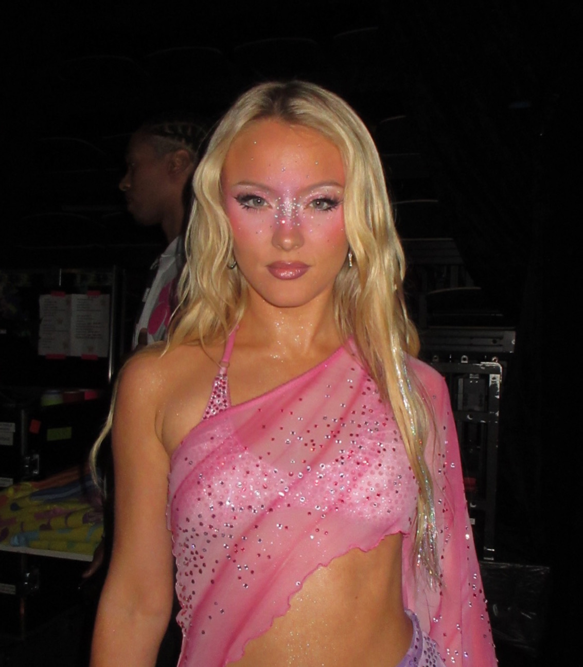 Zara Larsson looks straight at the camera backstage, long blonde hair down, glitter across her cheeks and brows, in a sheer pink crystal-studded top, with flight cases in the dark behind her.
