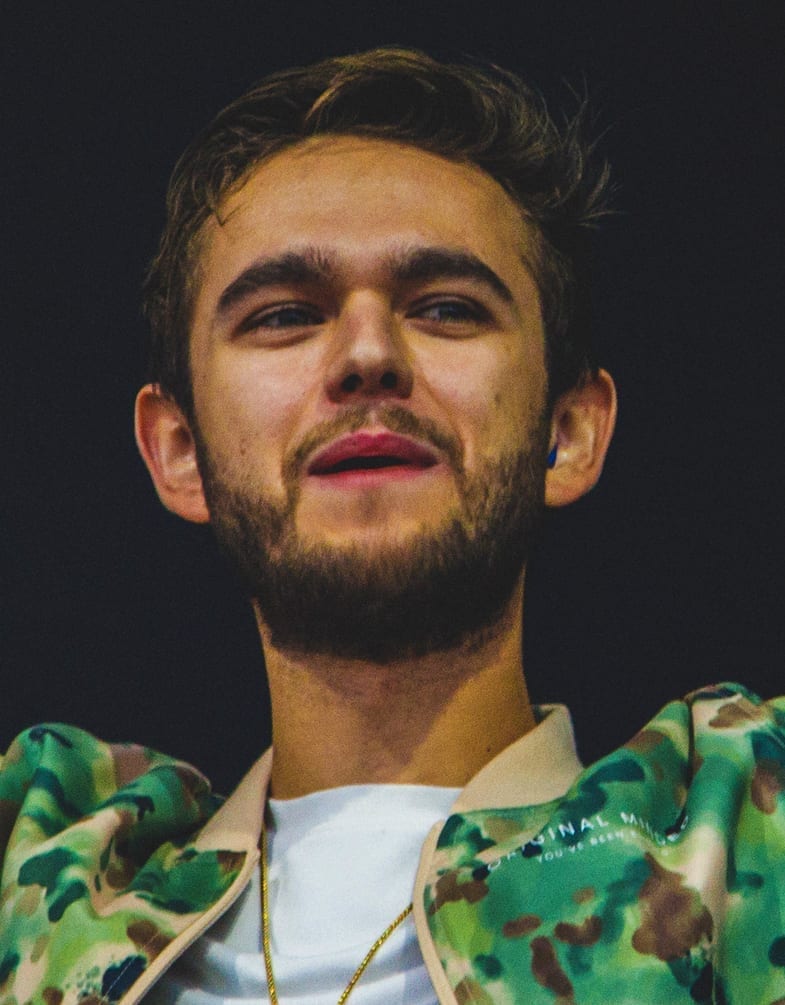 Zedd, wearing a camouflage-patterned jacket and gold chain, looks upward with earbuds visible.