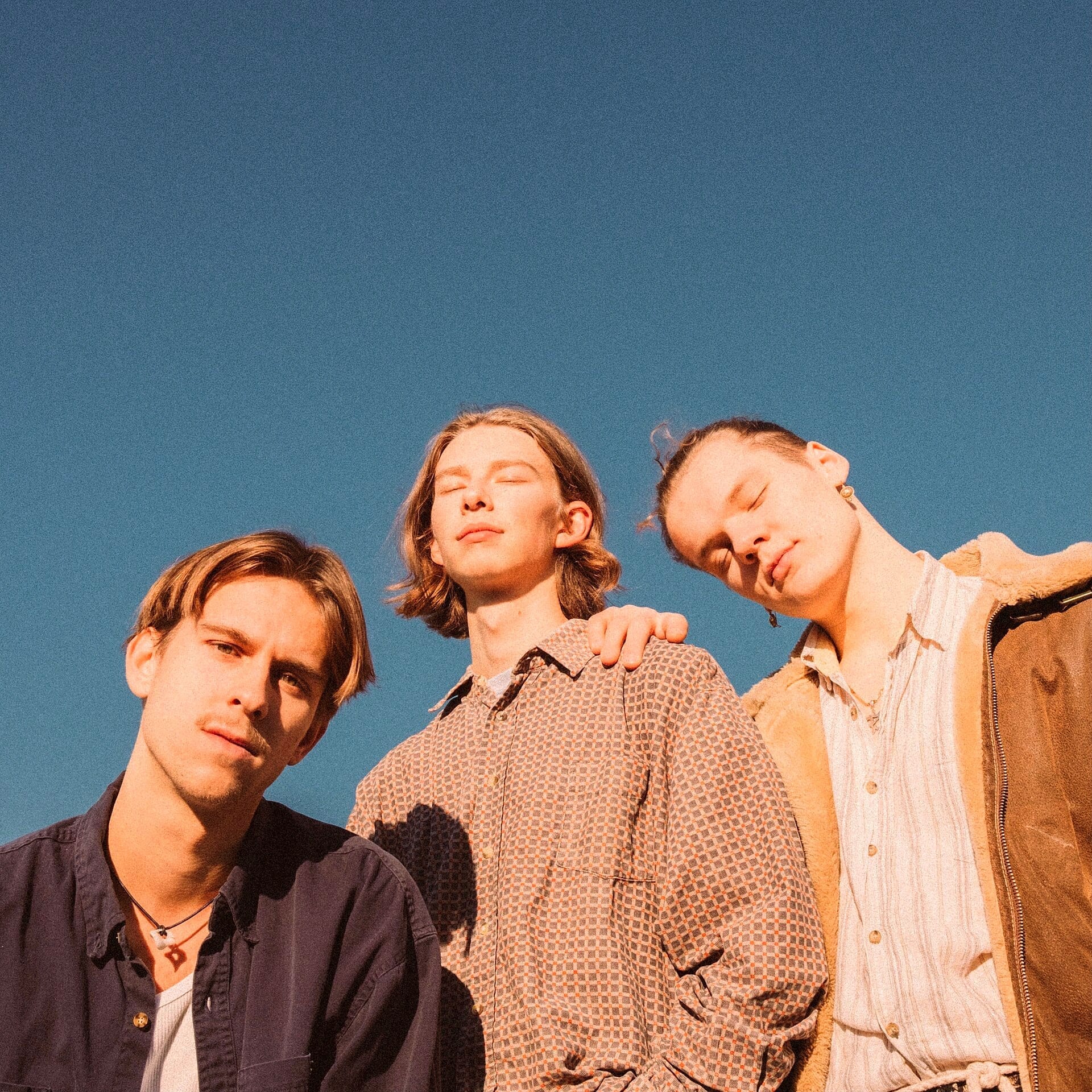 The three members of Zimmer90 stand close together outdoors against a clear blue sky, backlit by warm sunlight, wearing patterned button-up shirts and a shearling jacket.